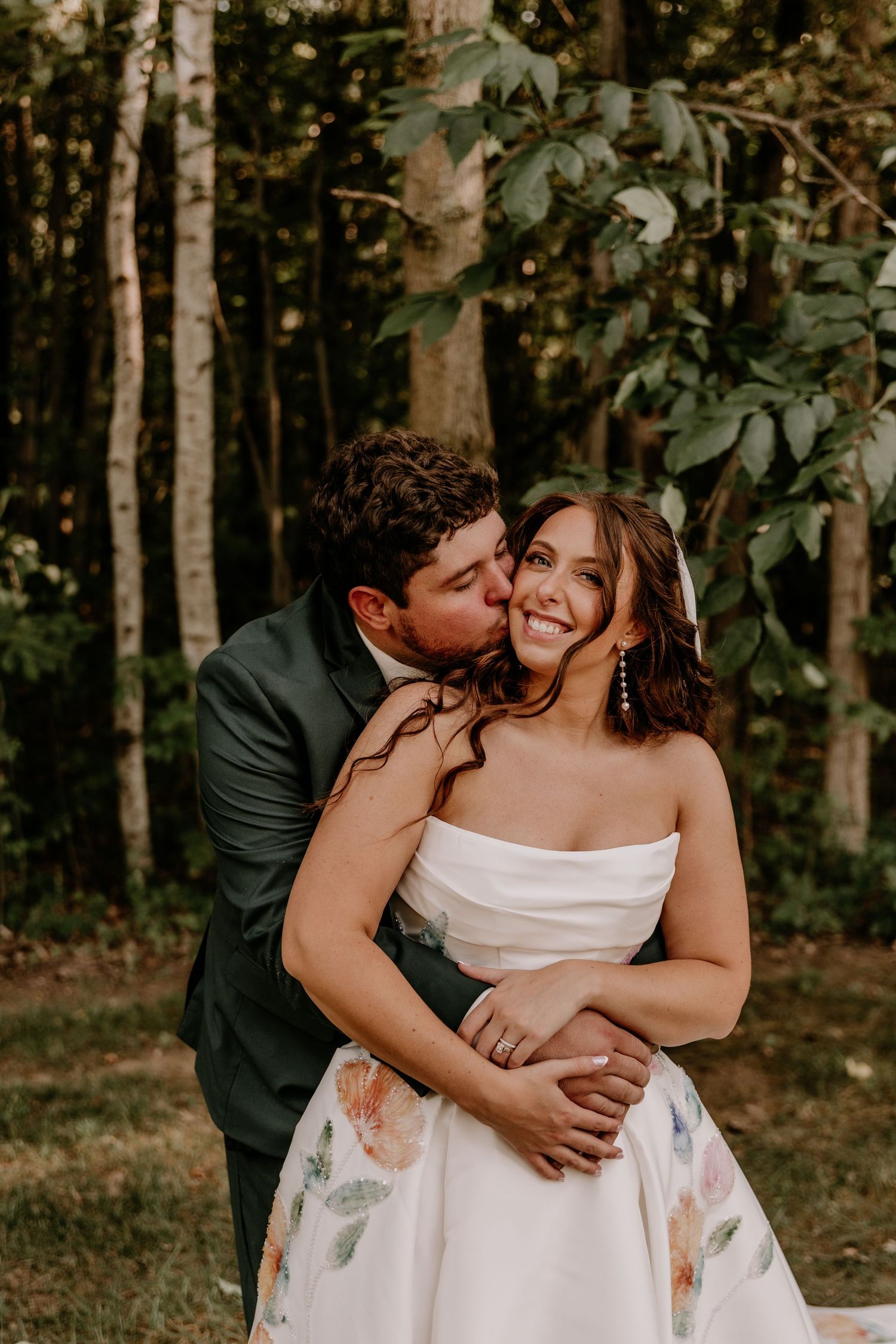 Groom in dark suit kisses bride on cheek as she smiles, standing outdoors in lush green forest setting.