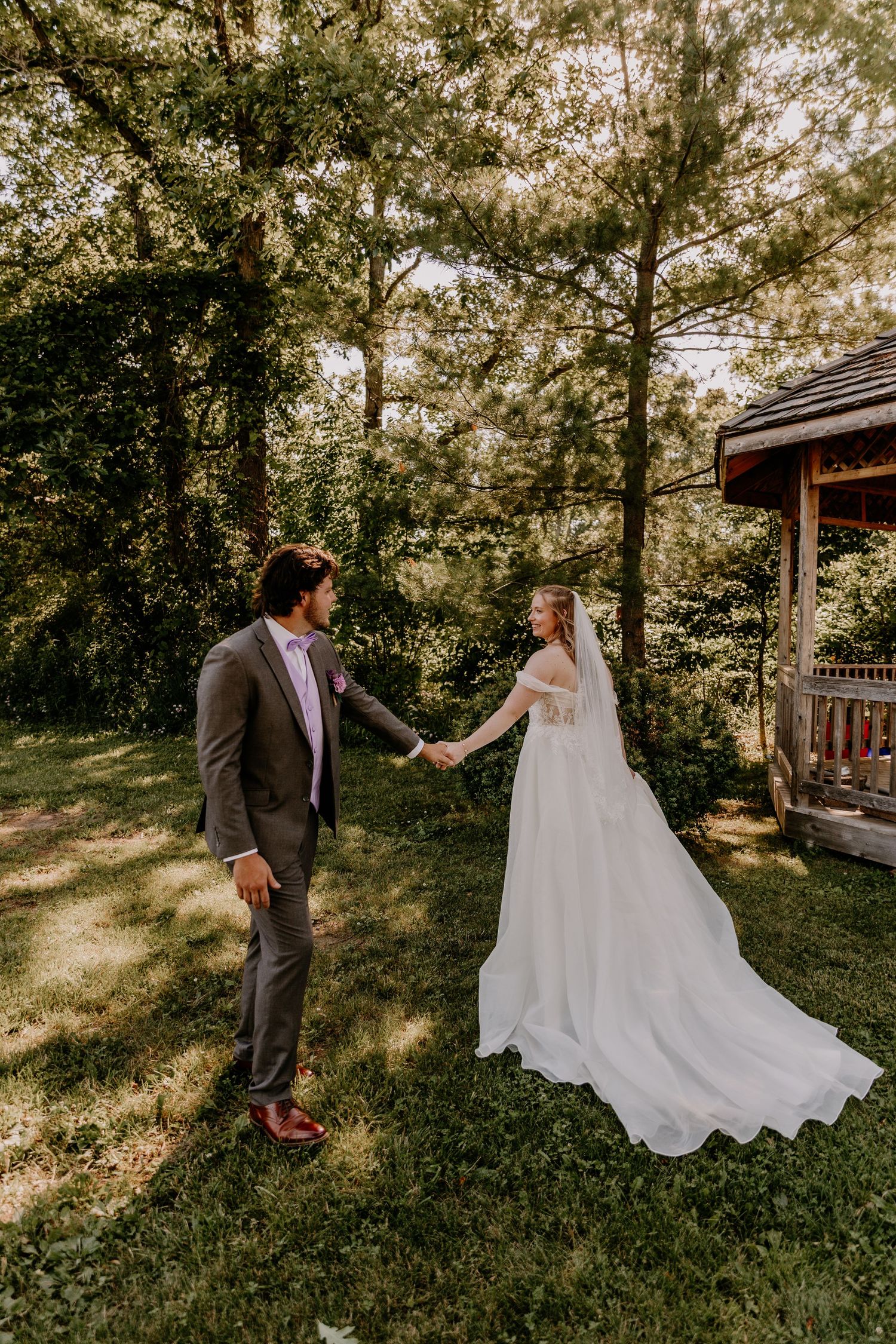 Bride and groom hold hands in lush garden setting, bride wearing flowing white gown with veil near rustic gazebo.