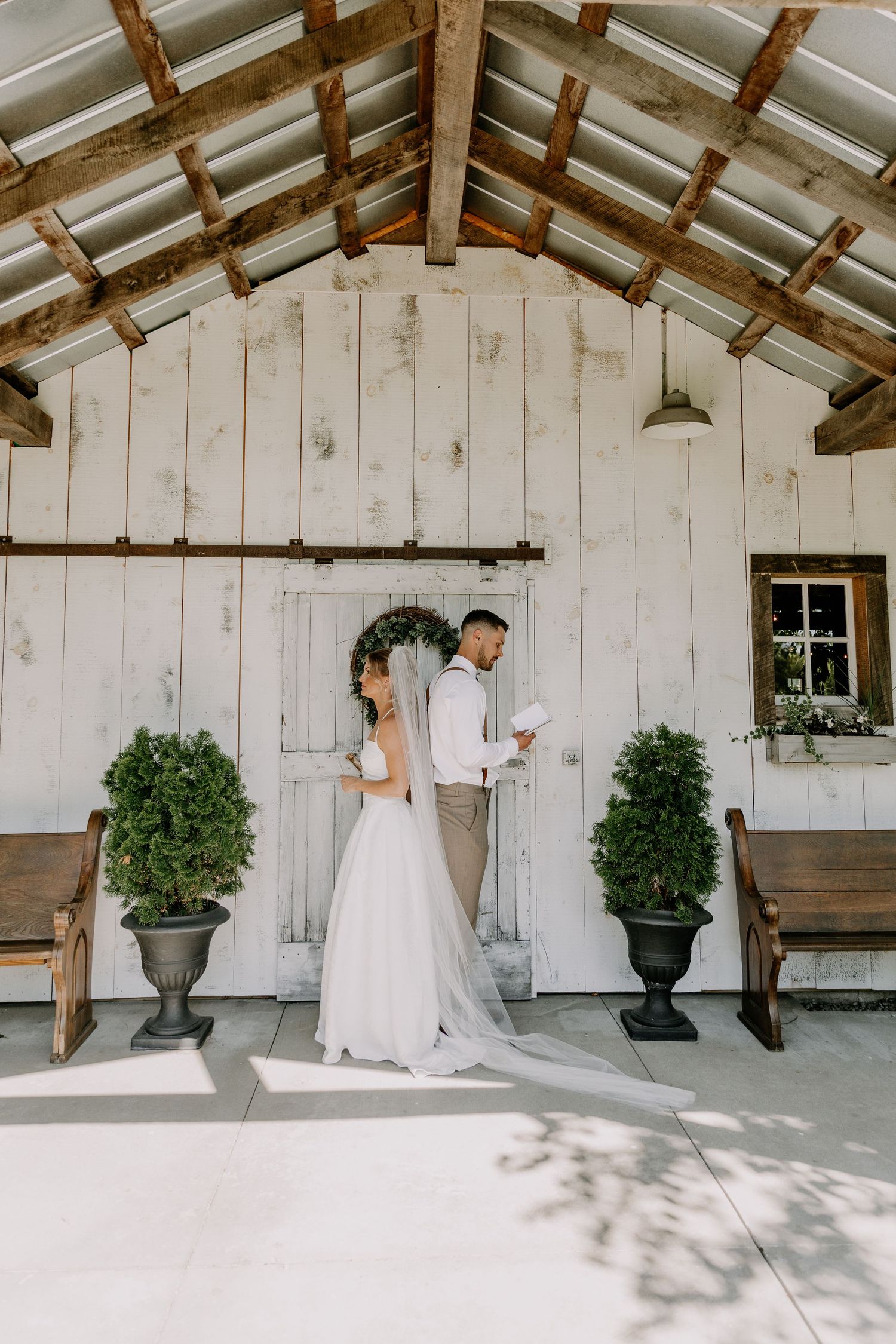 Bride and groom share vows outside a rustic white barn chapel with wood beams, potted topiaries, and vintage benches.