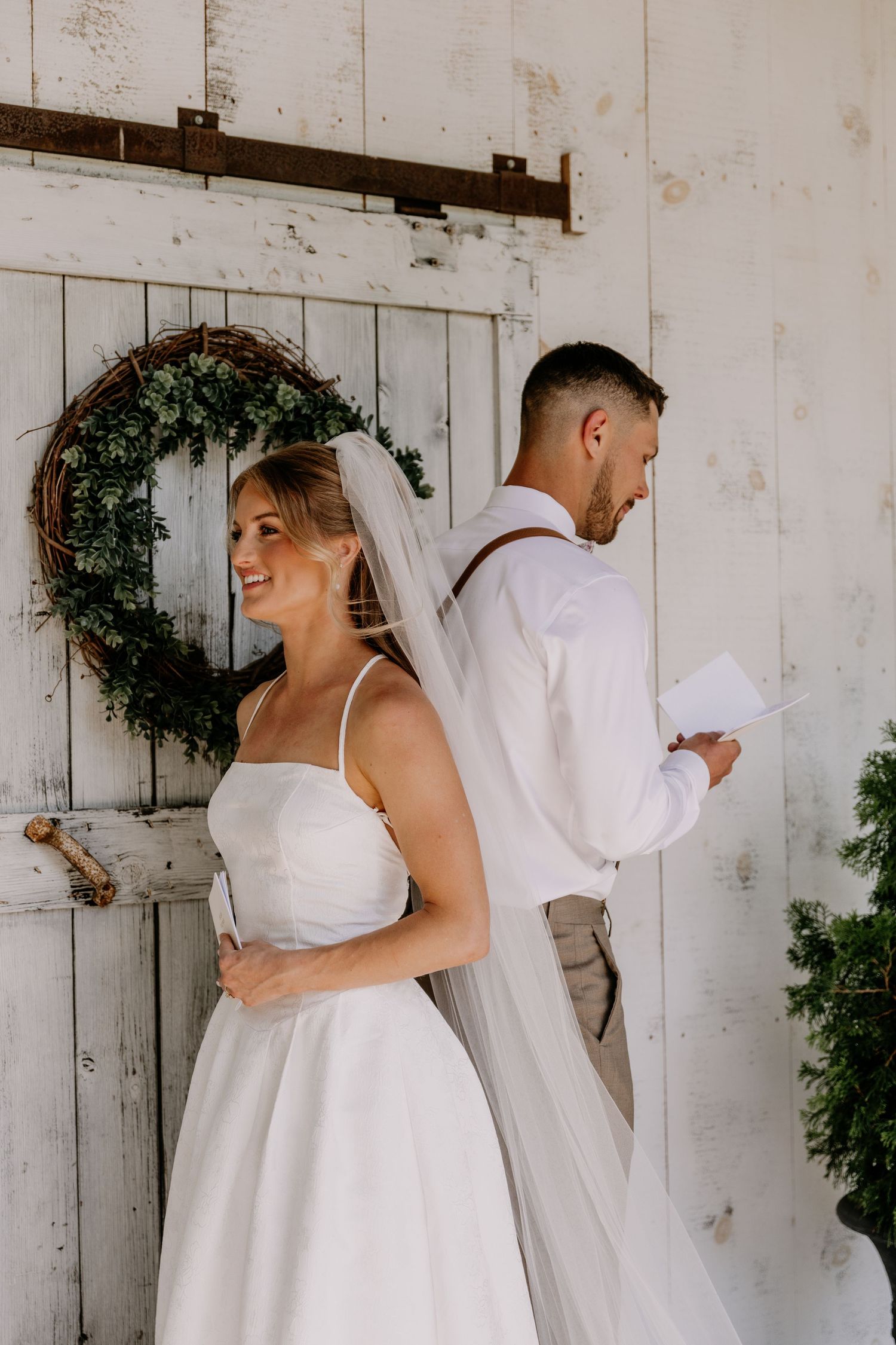 Smiling bride in a white gown and veil stands back-to-back with groom reading vows by a rustic barn door with wreath.