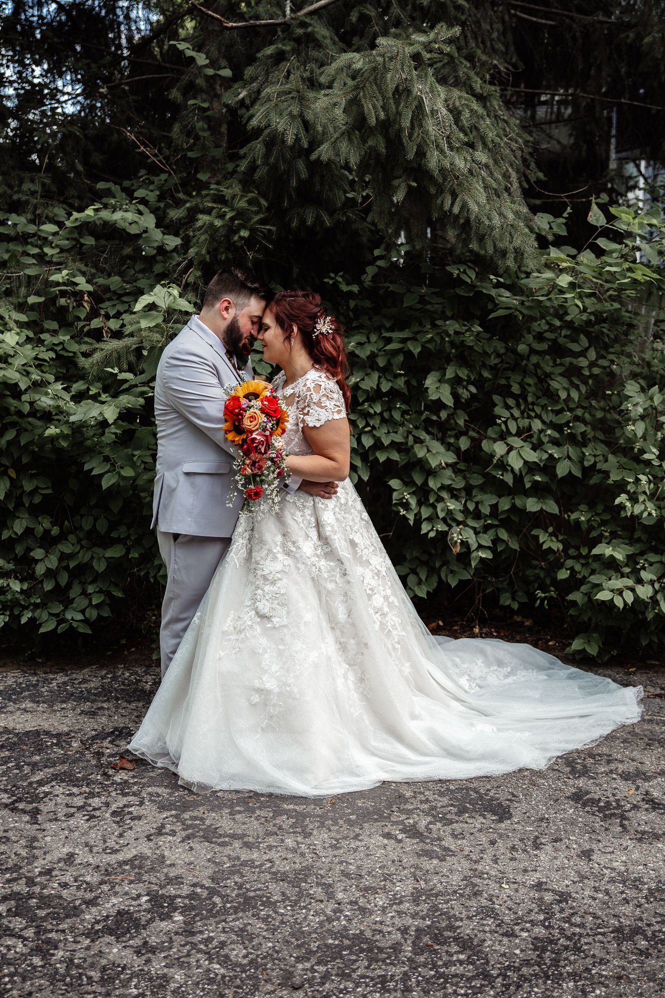A wedding couple embraces in front of lush greenery while wearing a grey suit and white lace gown holding vibrant flowers.