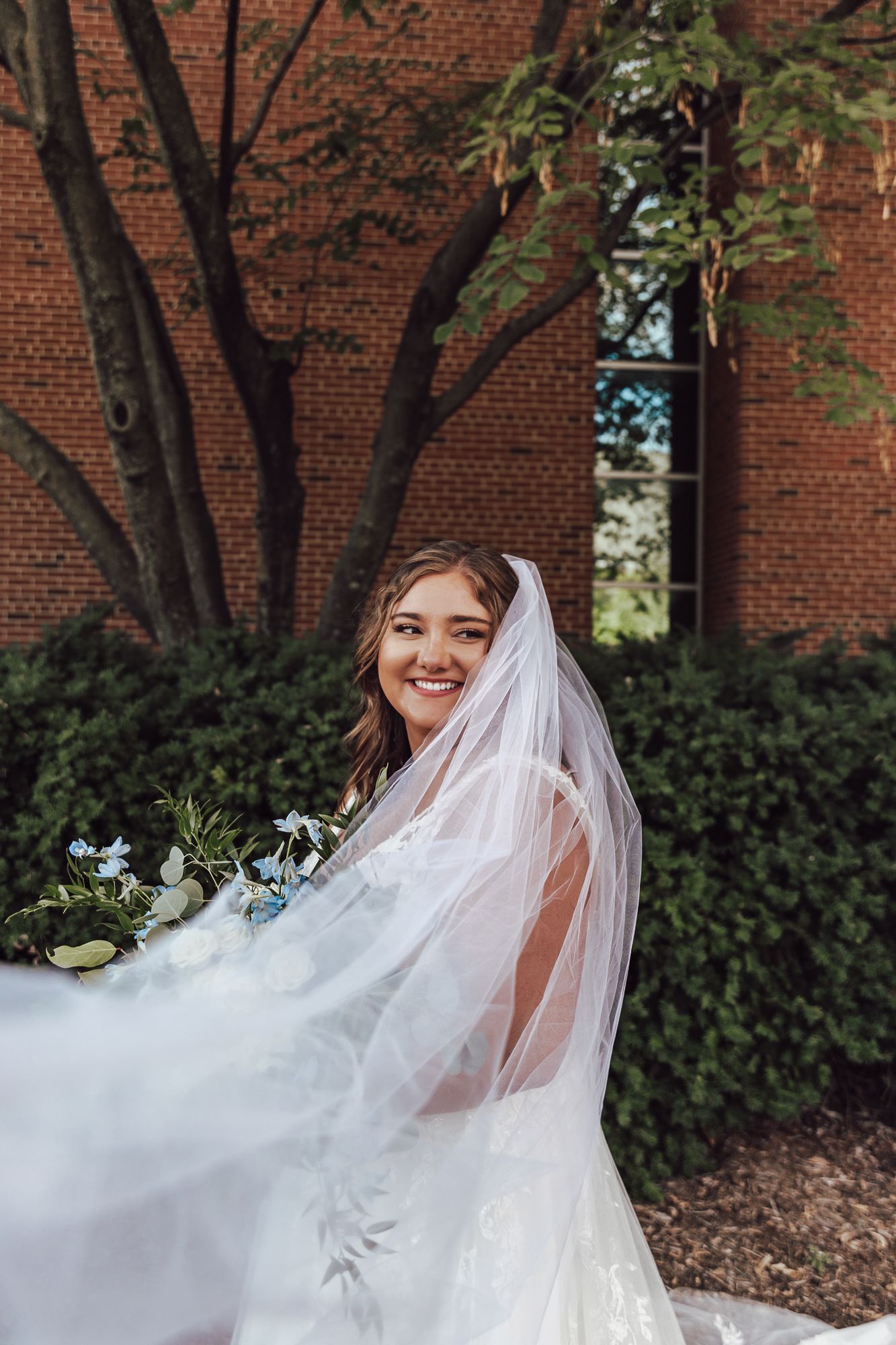Bride with flowing veil smiles while holding white and blue bouquet against brick church wall.