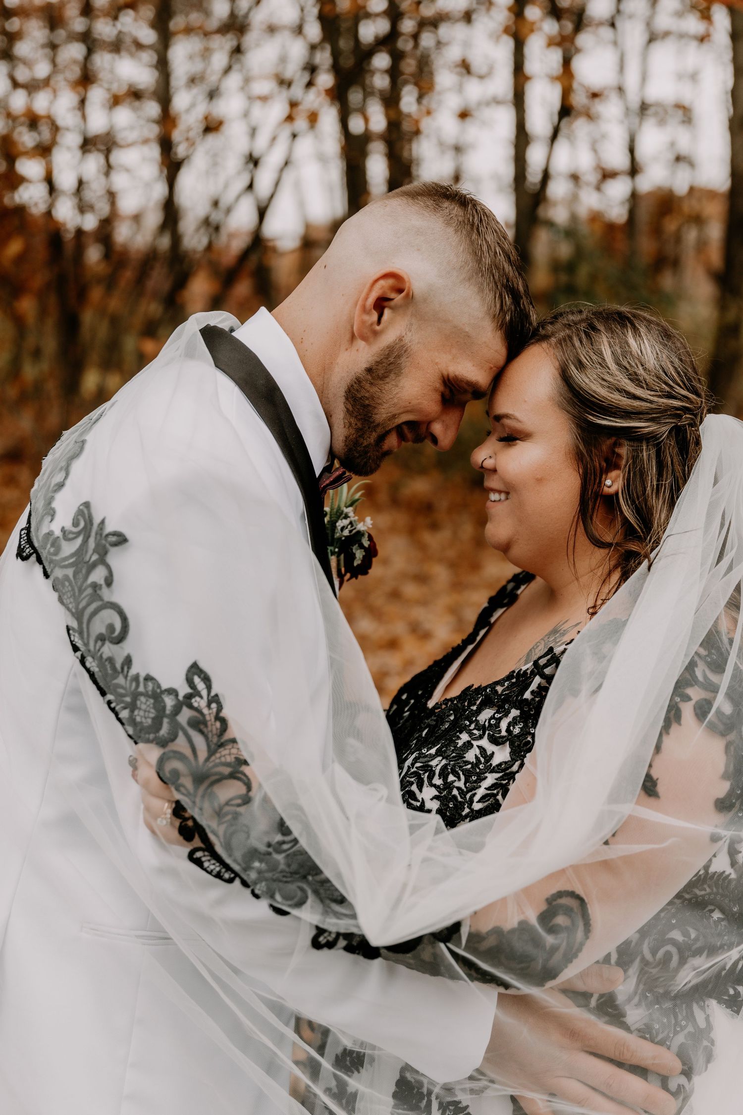Intimate moment between newlyweds in autumn forest with bride wearing detailed white dress and veil.