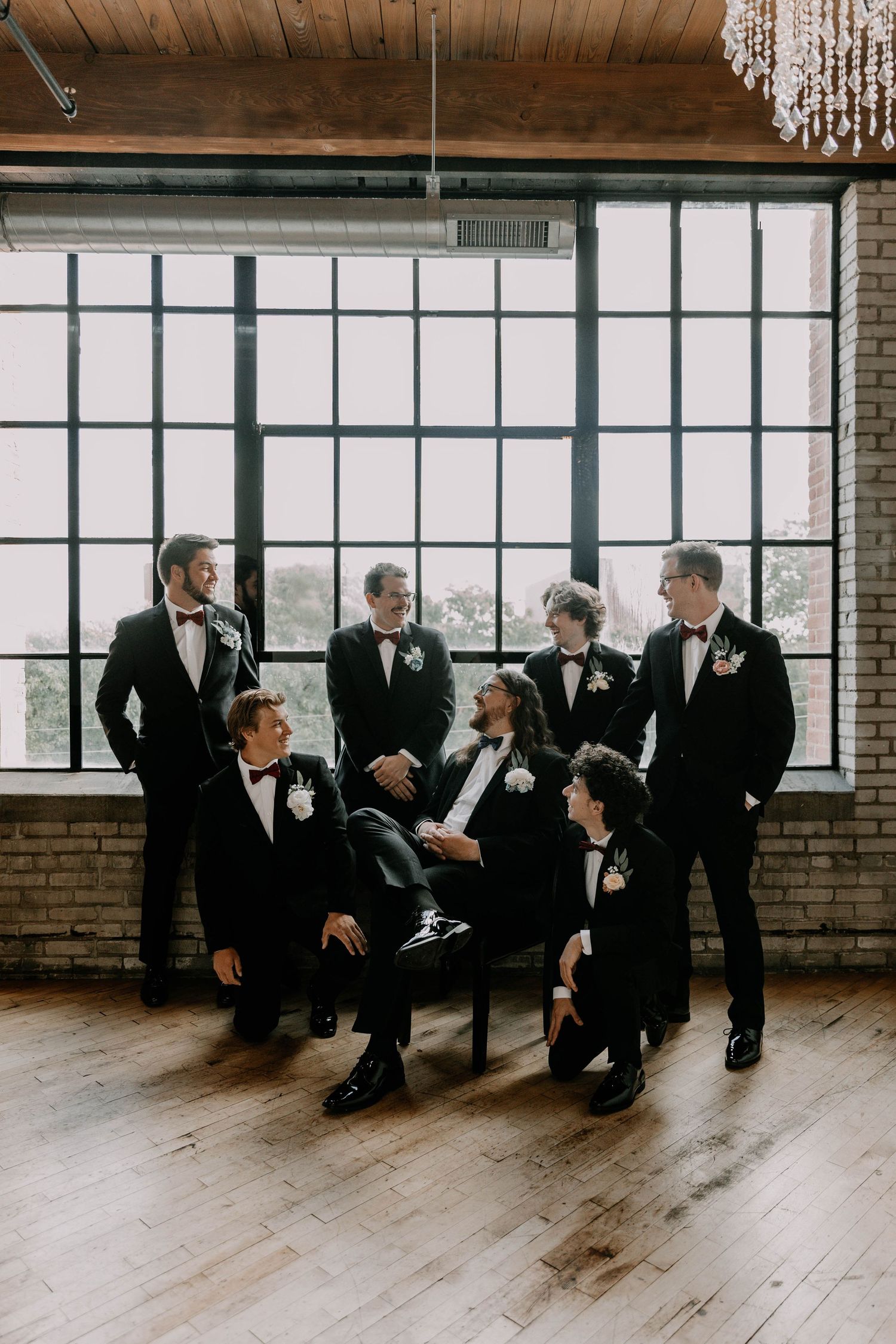 A group in black tuxedos poses together in an industrial loft space with large windows.