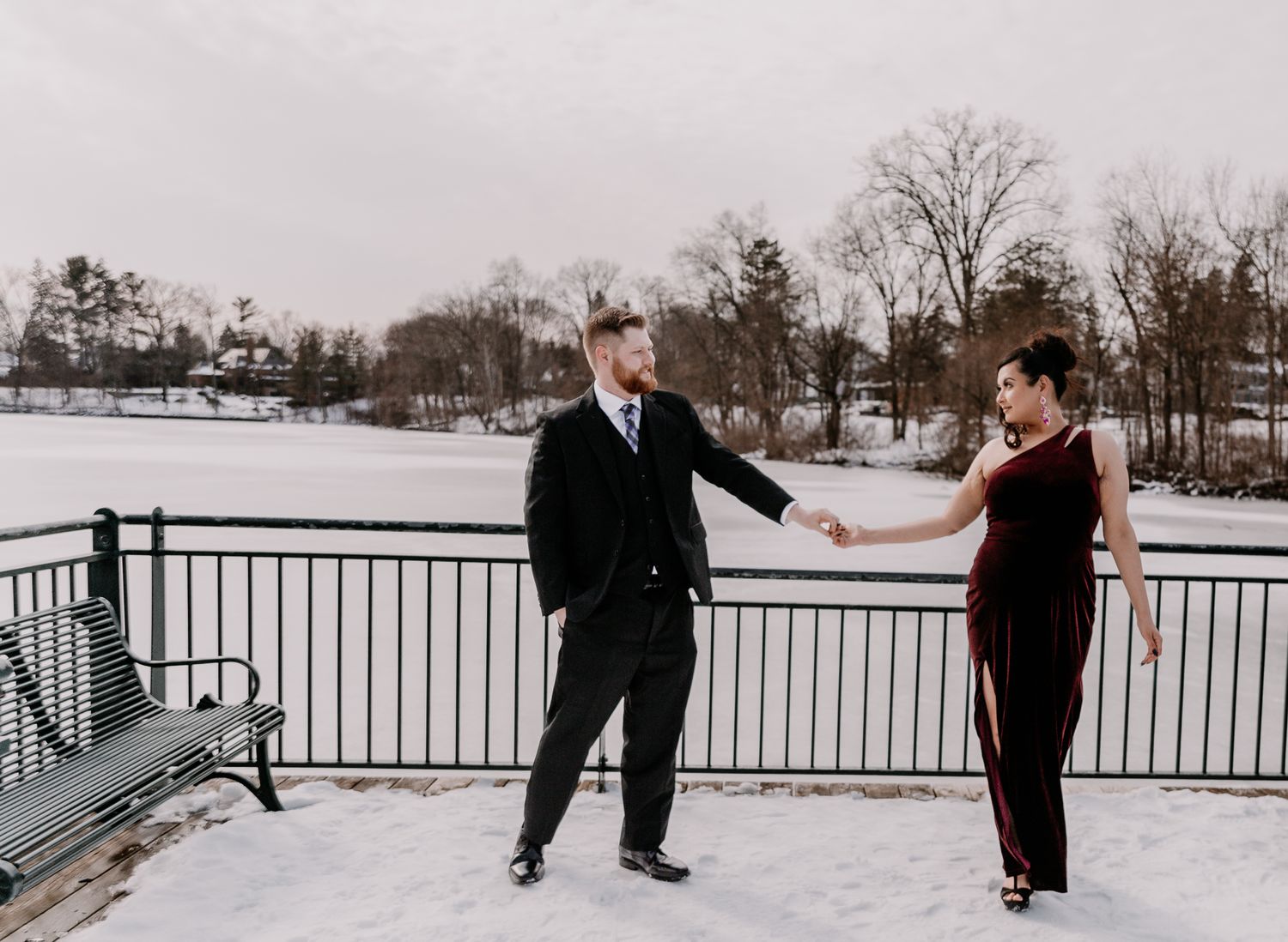 A couple stands on a snow-covered bridge railing sharing a playful moment together.