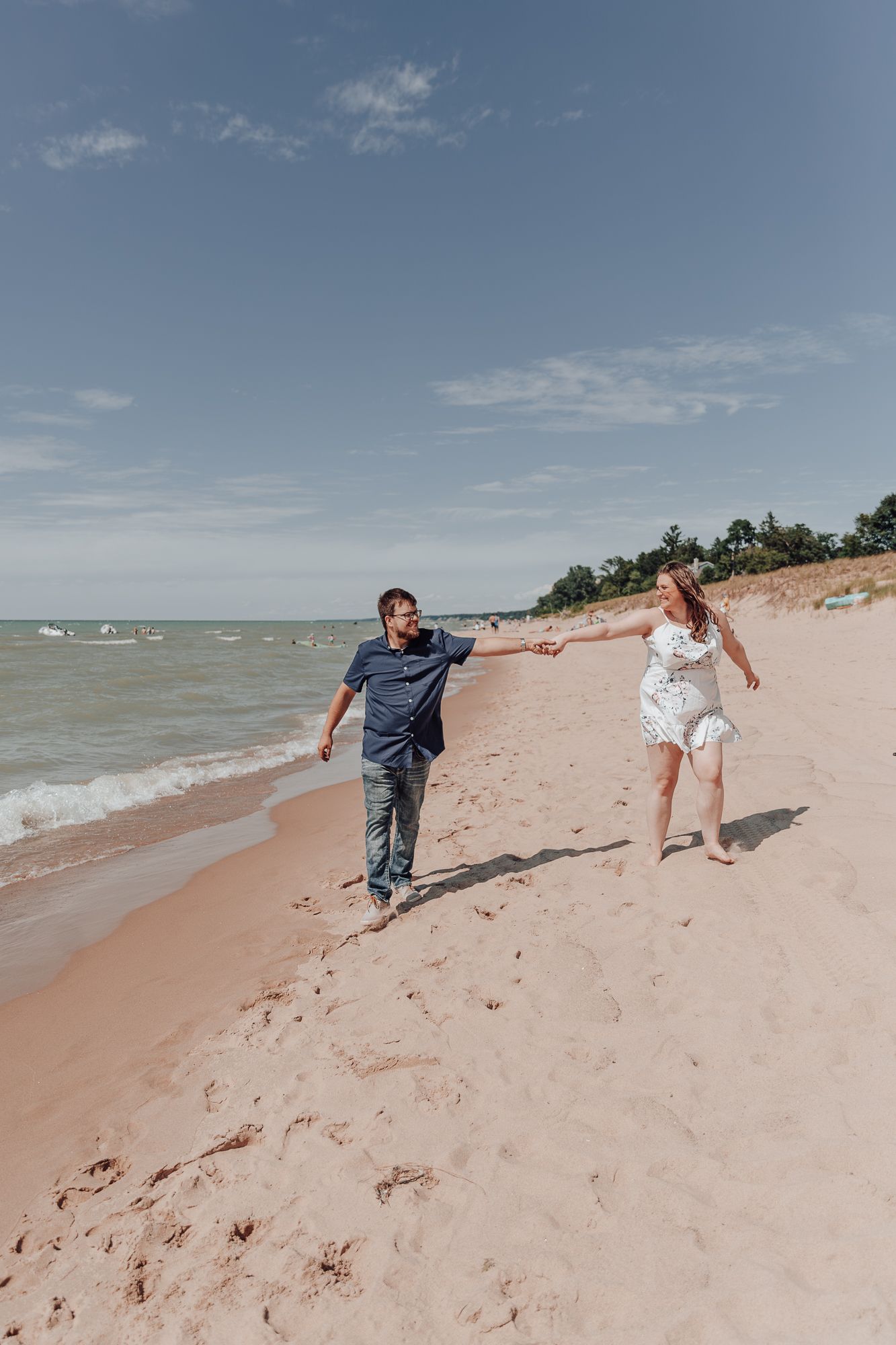 Playful couple running along shoreline with waves and sandy beach dunes in background.