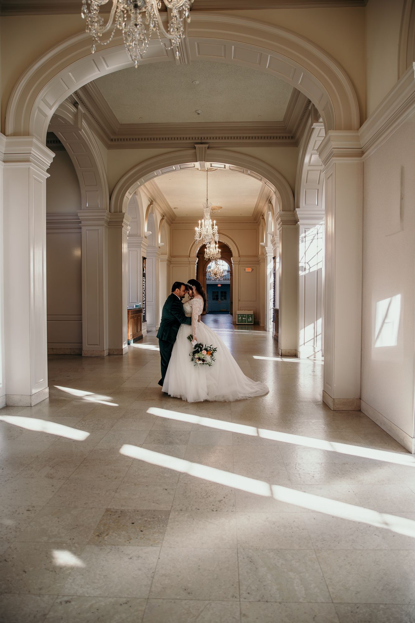 A couple sharing an intimate moment in a grand hallway with ornate architectural details and dramatic lighting.