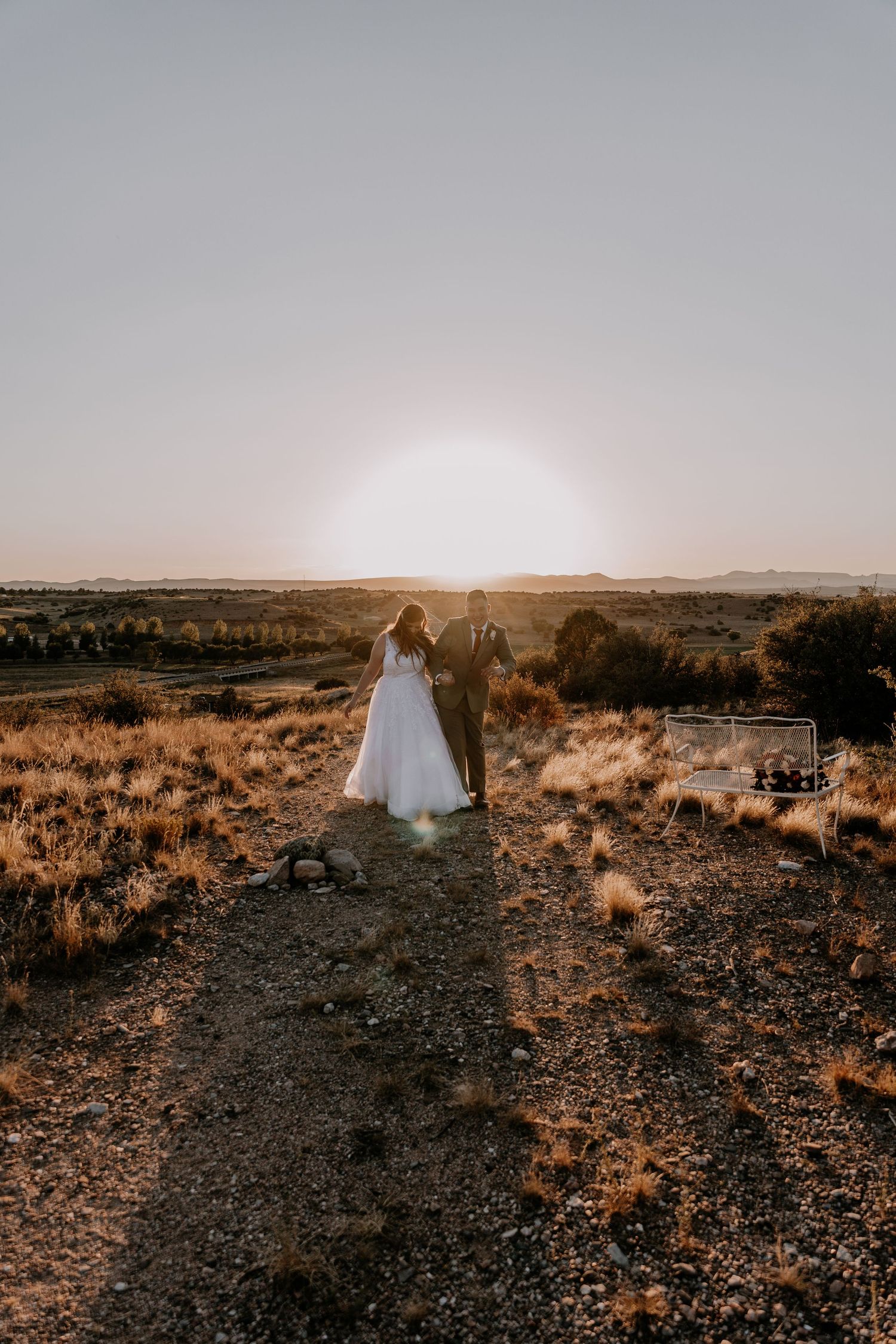 Wide landscape shot of wedding couple in desert terrain with mountains and sunset in background.