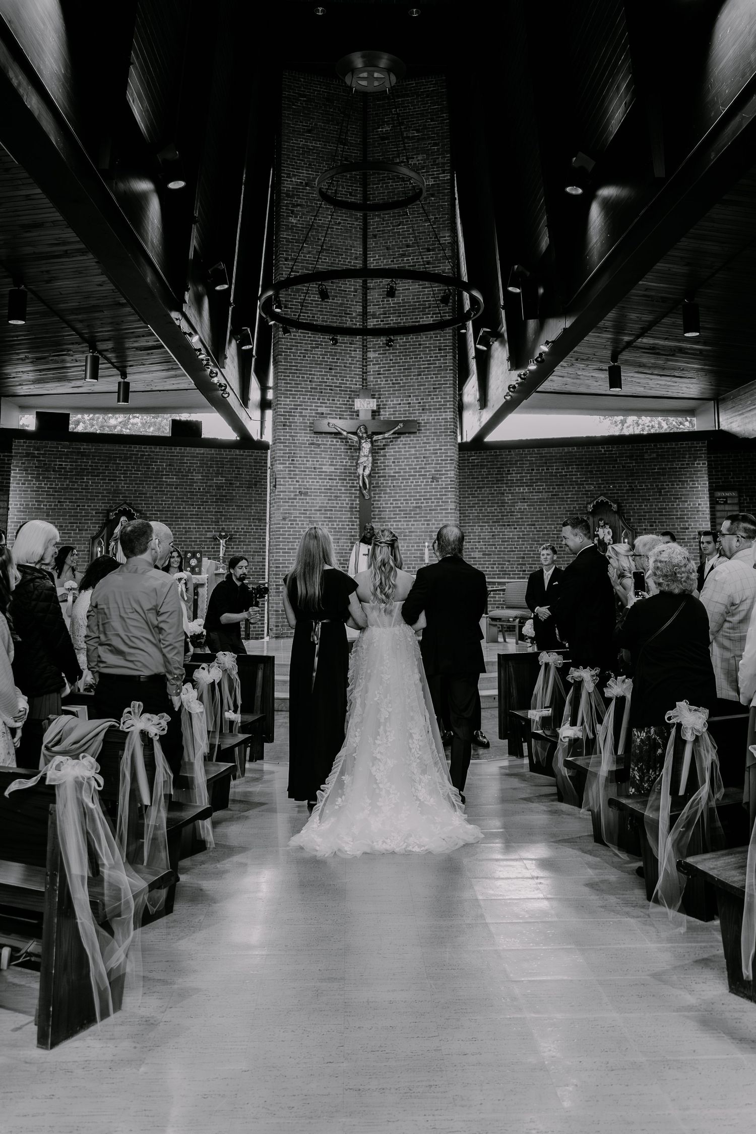 Black and white photo of a bride walking down the church aisle toward a crucifix, flanked by guests in pews.