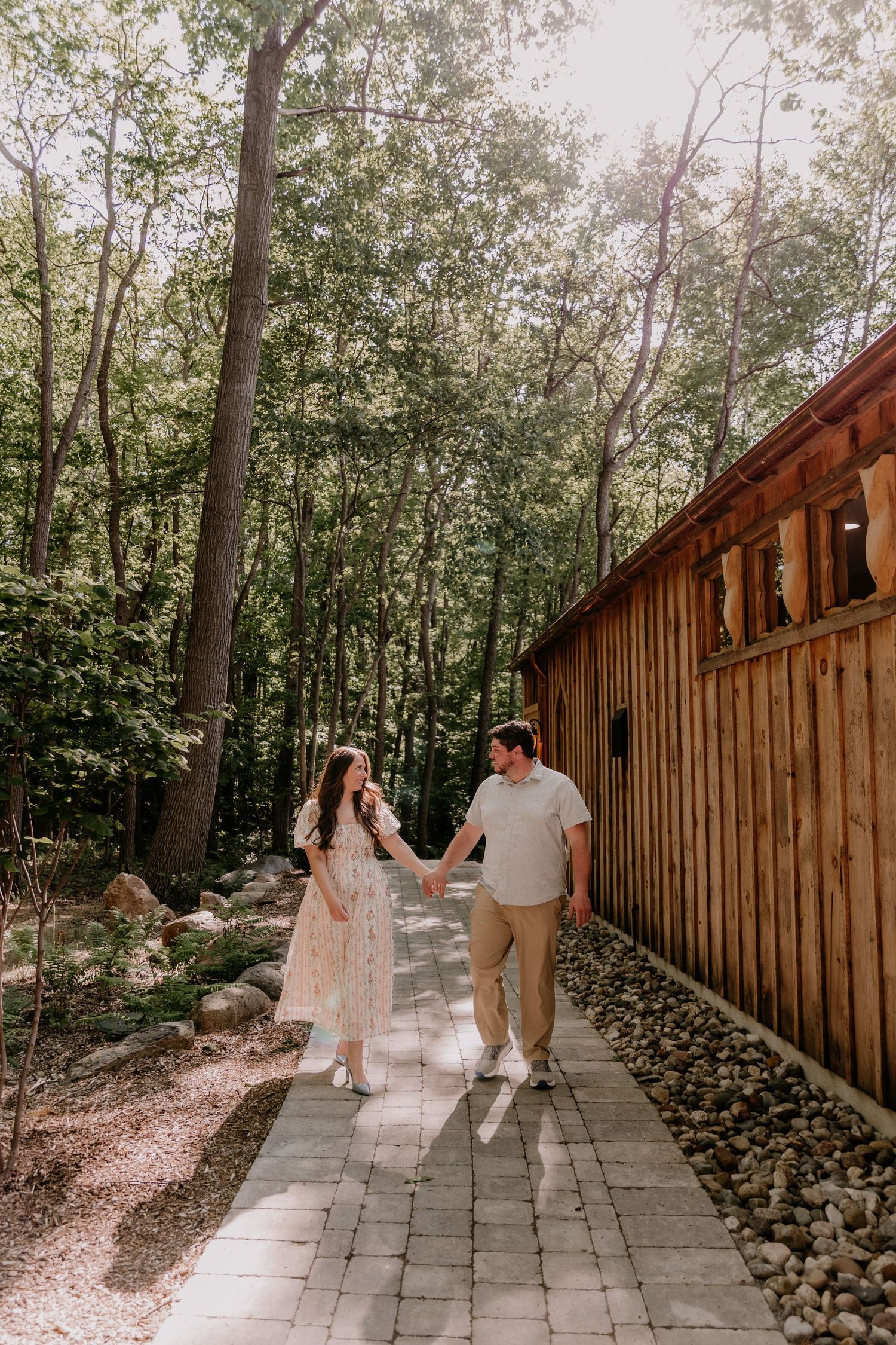 Couple holding hands walking along a stone path beside a wooden barn surrounded by lush green forest trees.