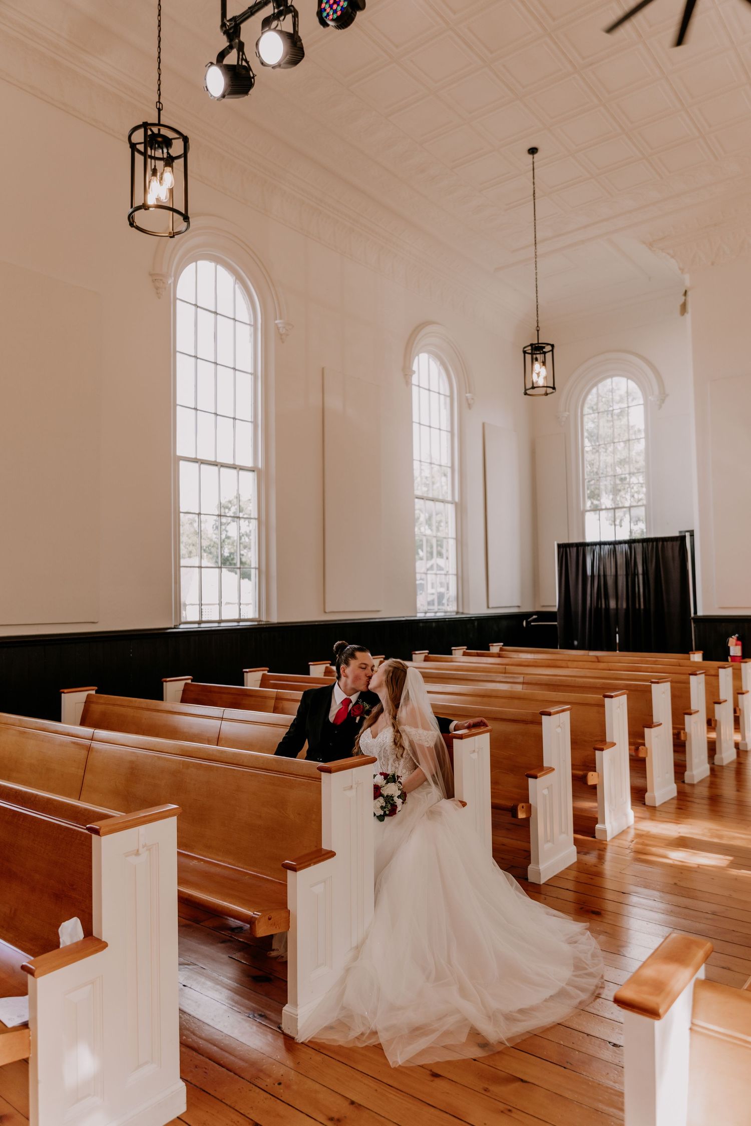 A bride and groom share a kiss in a sunlit white church with wooden pews on their wedding day.