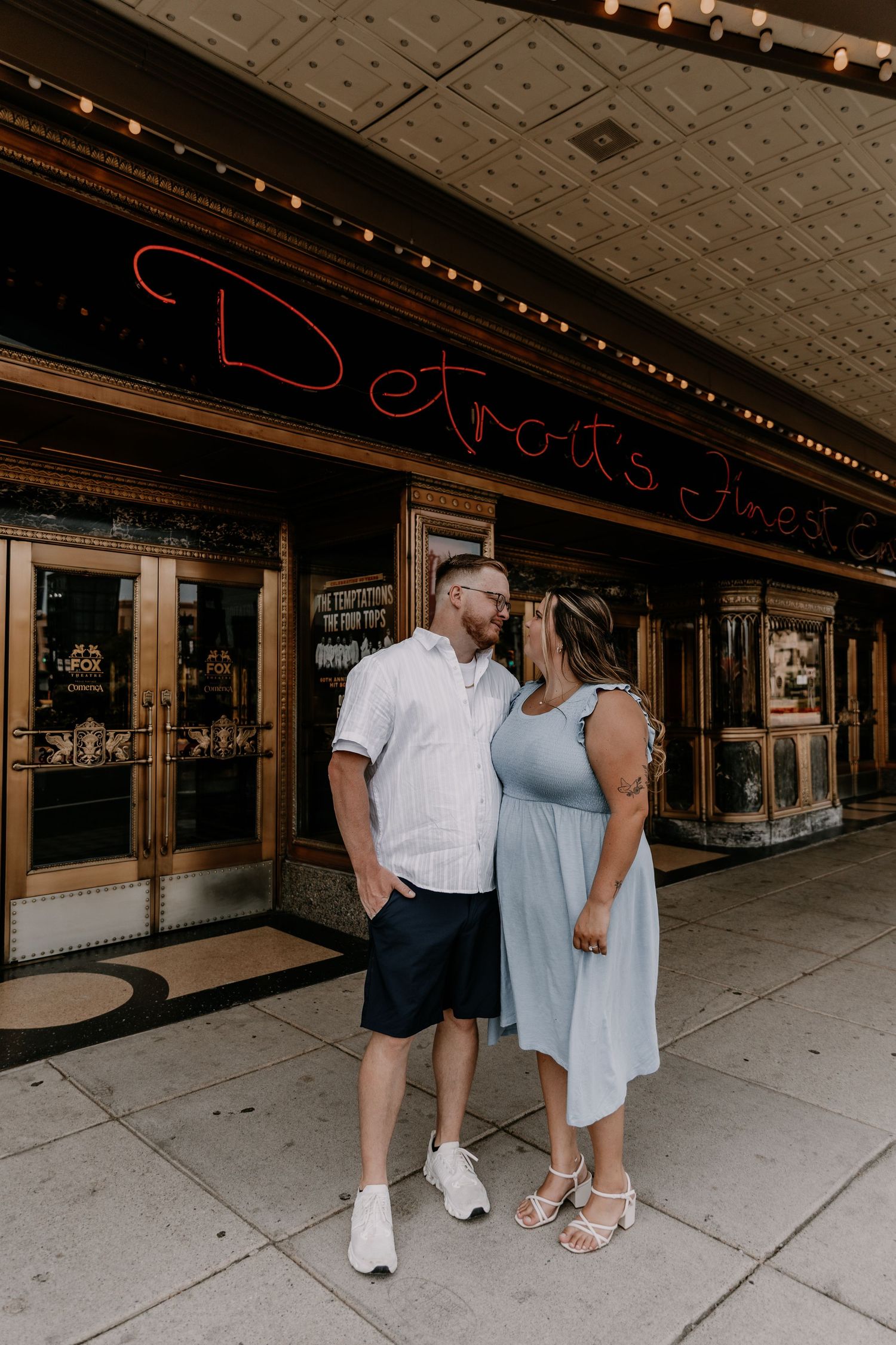 Couple walking together and embracing along a historic storefront with a decorative tin ceiling and string lights overhead.