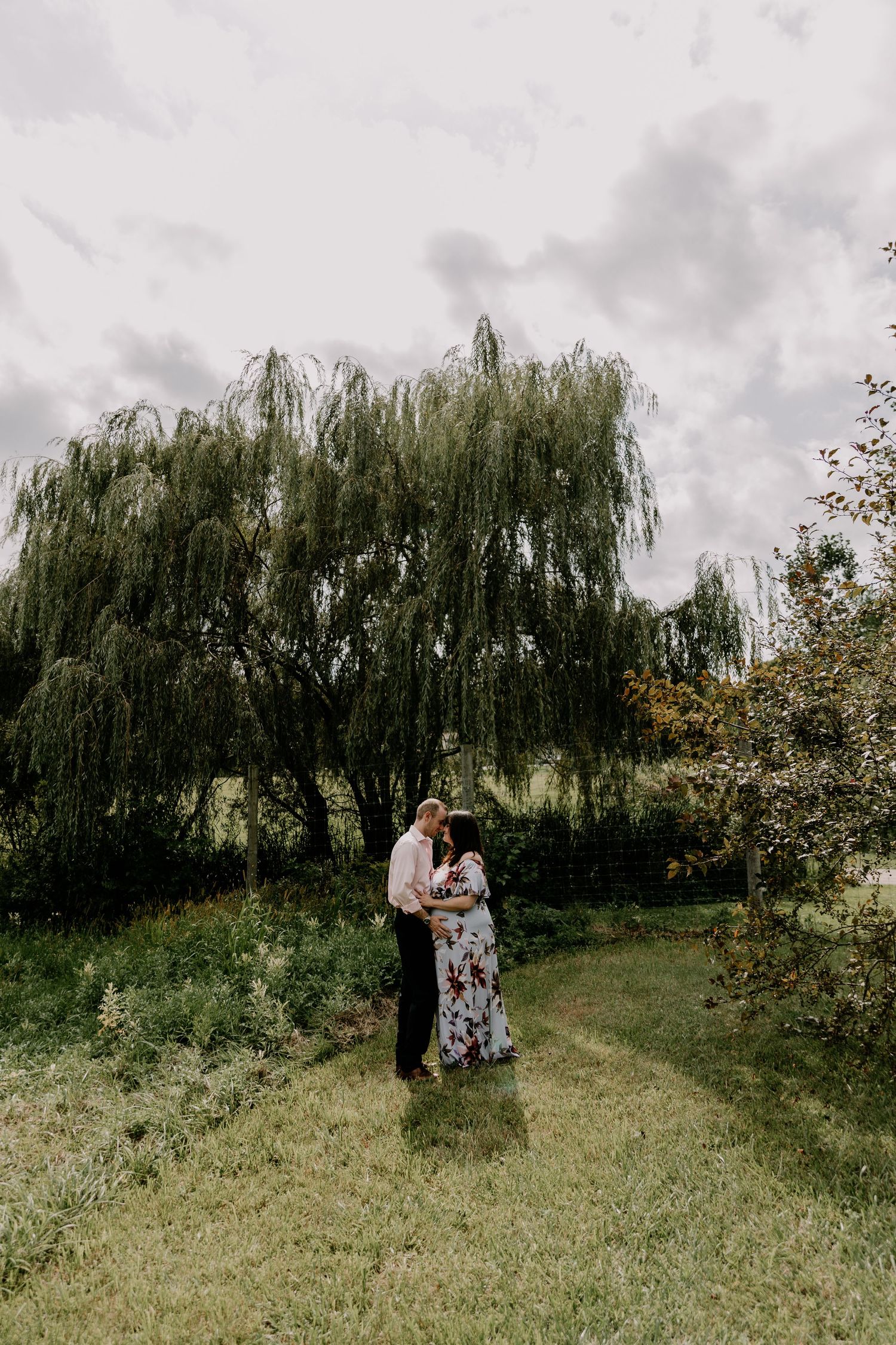 A couple embraces on a grassy hillside under dramatic weeping willow trees on an overcast day.