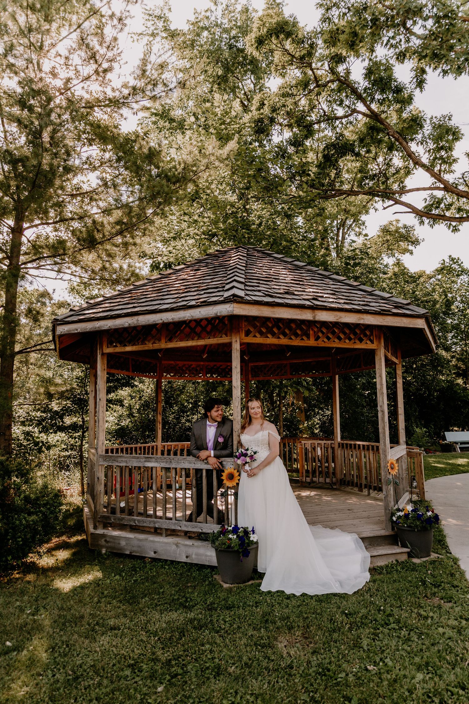 Bride and groom posing together in front of a rustic wooden gazebo surrounded by lush green trees.