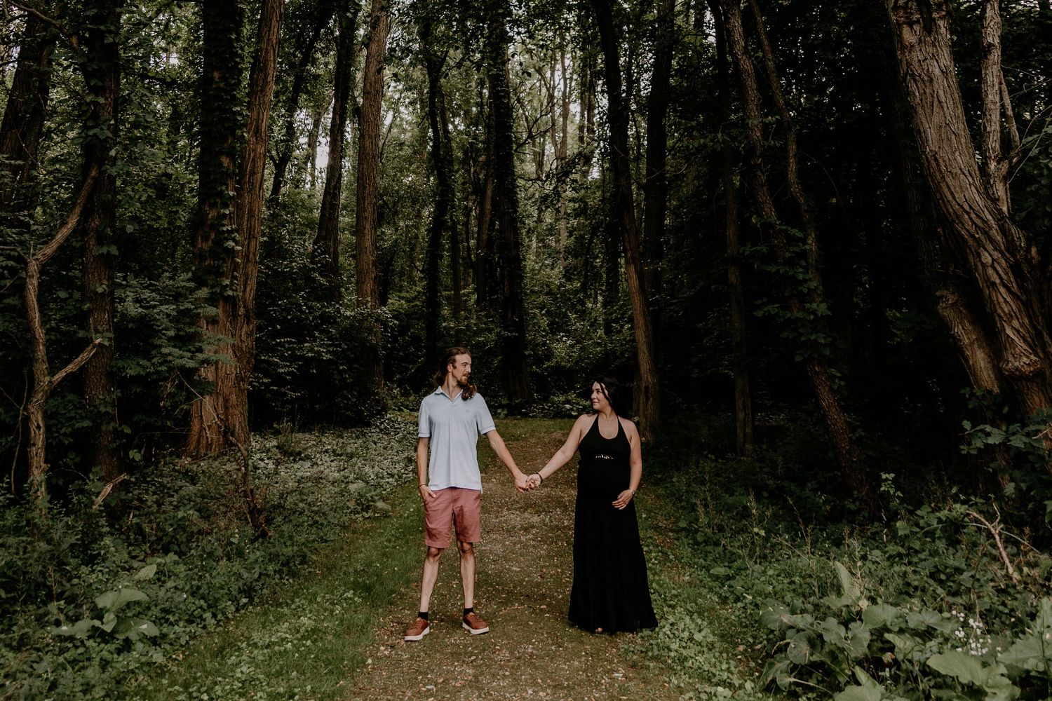 Couple holding hands on a forest path surrounded by tall trees and lush greenery in an outdoor engagement photo.