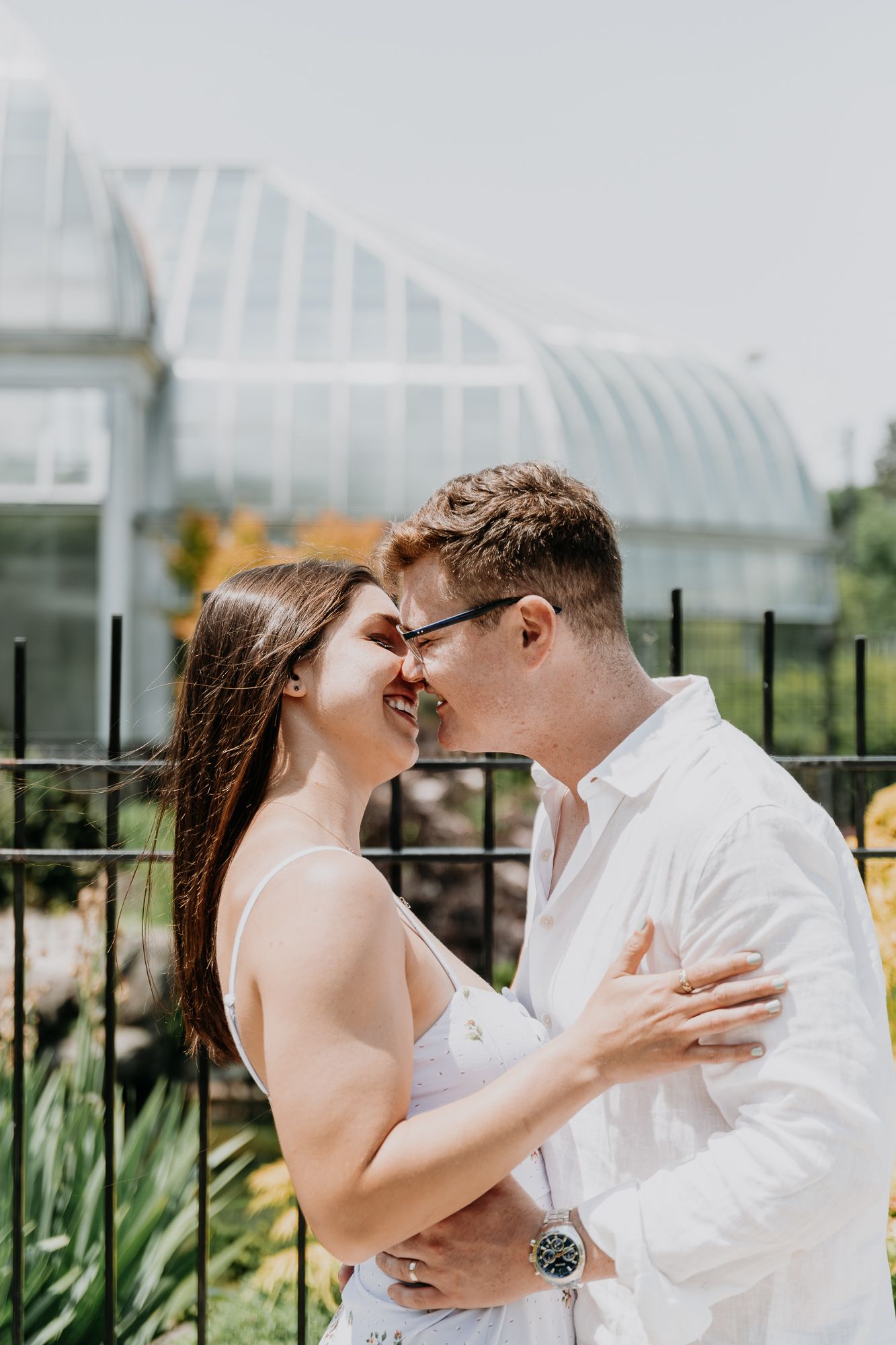 A couple embraces near a greenhouse garden with glass panels visible in the background.