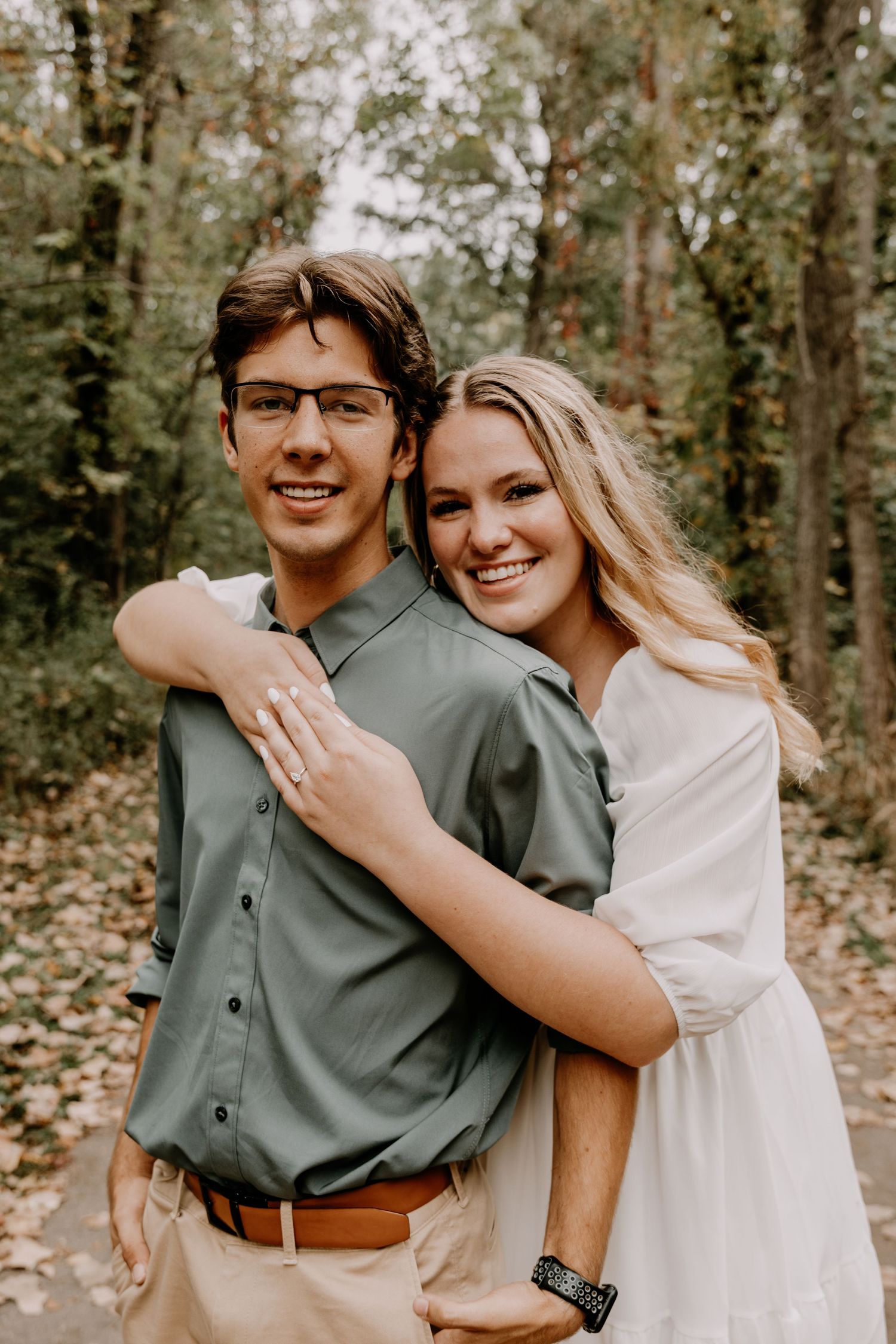 A couple poses for an engagement photo in a wooded setting, showing off her ring.