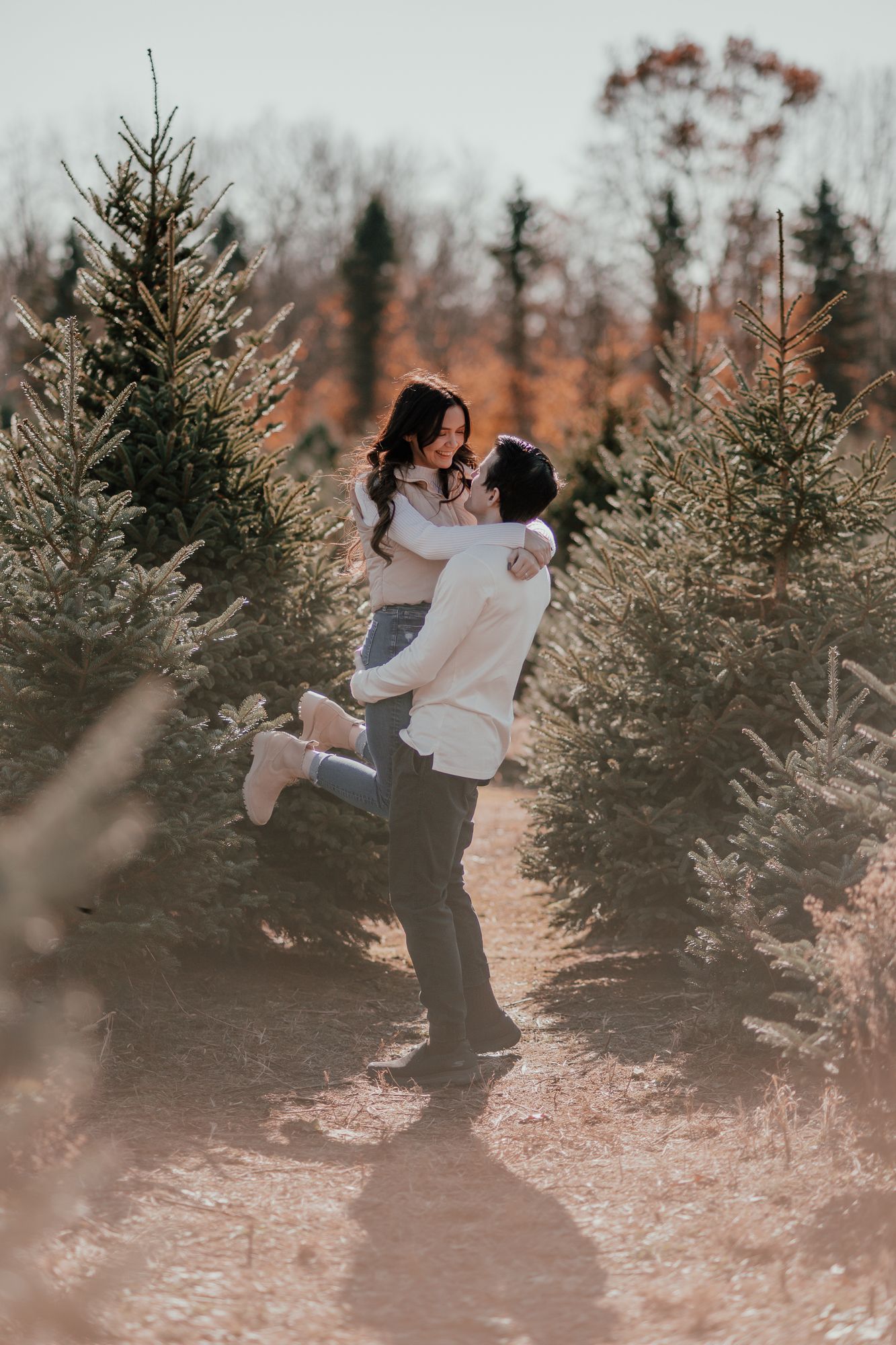 Man lifts partner in romantic embrace among Christmas trees during sunset engagement session.
