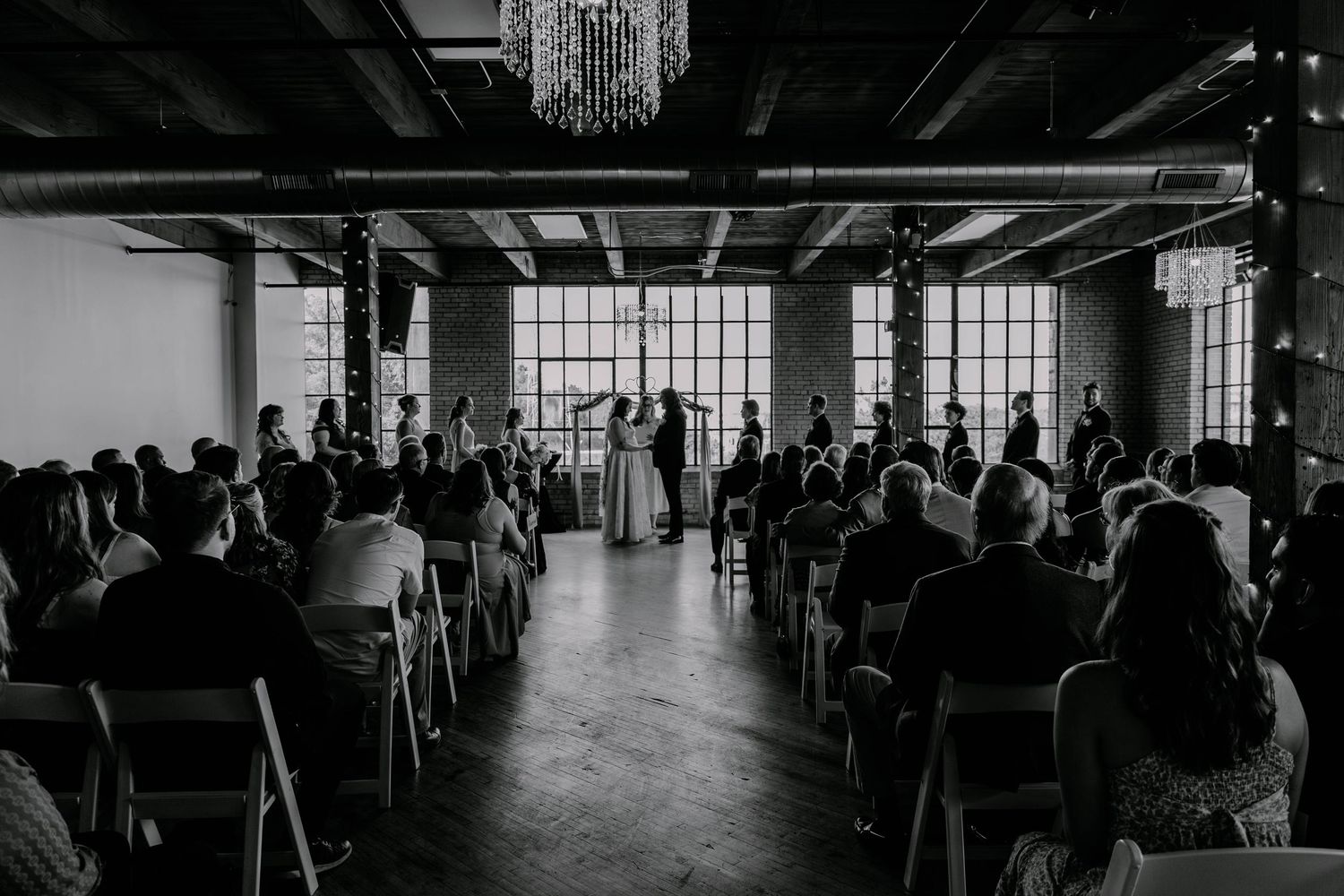 Black and white photo of wedding ceremony with guests seated in industrial venue with large windows.