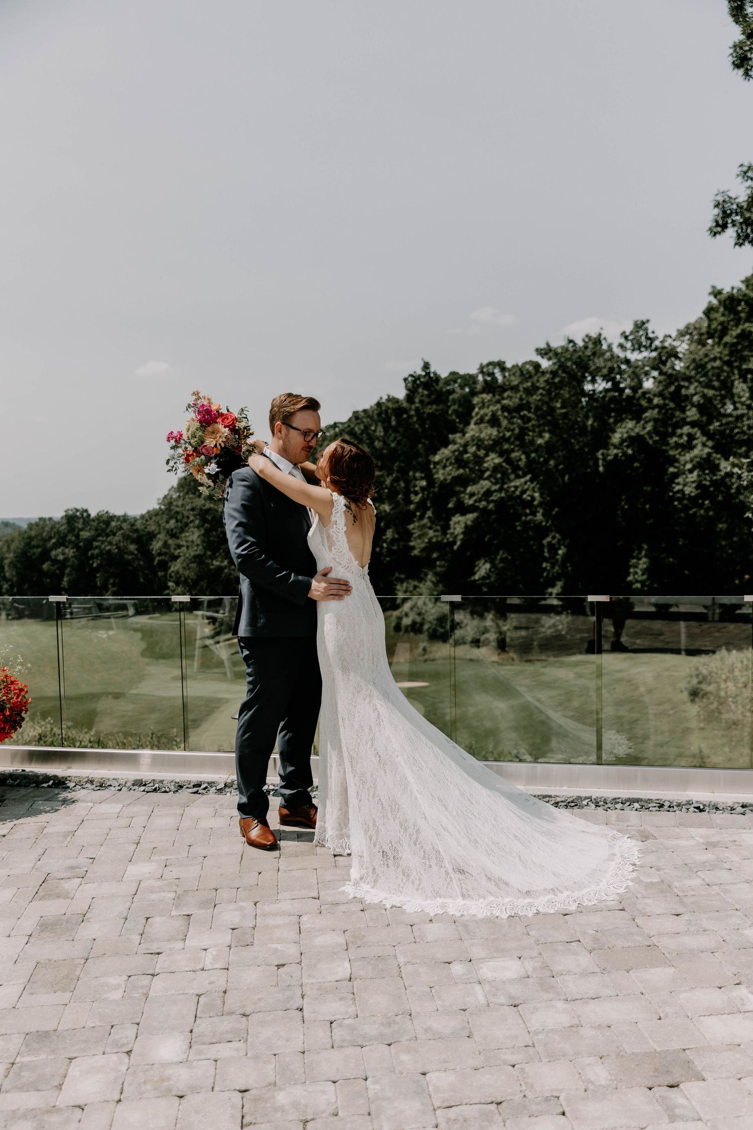 Romantic wedding couple embracing on stone patio with scenic countryside view through glass railing.