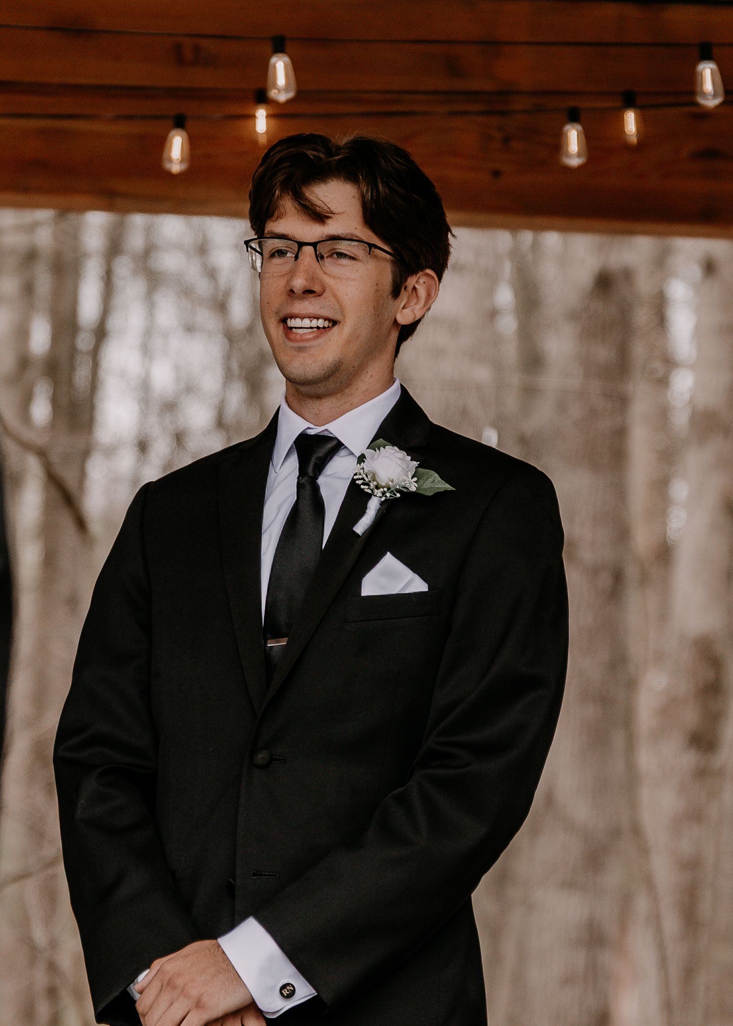 Groom in black suit with glasses smiling during wedding ceremony.