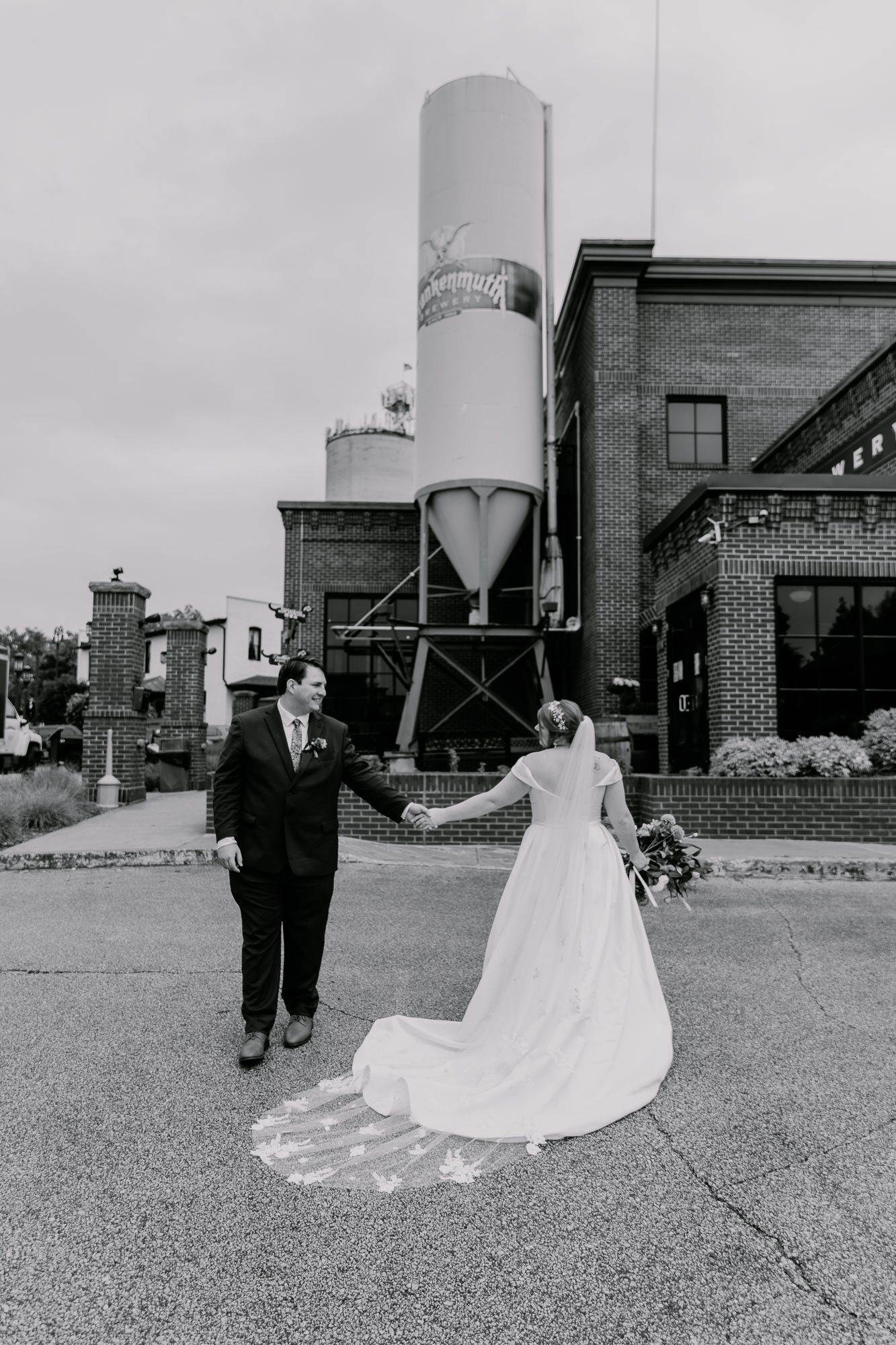 Newlyweds holding hands outside industrial building with large silos in black and white.