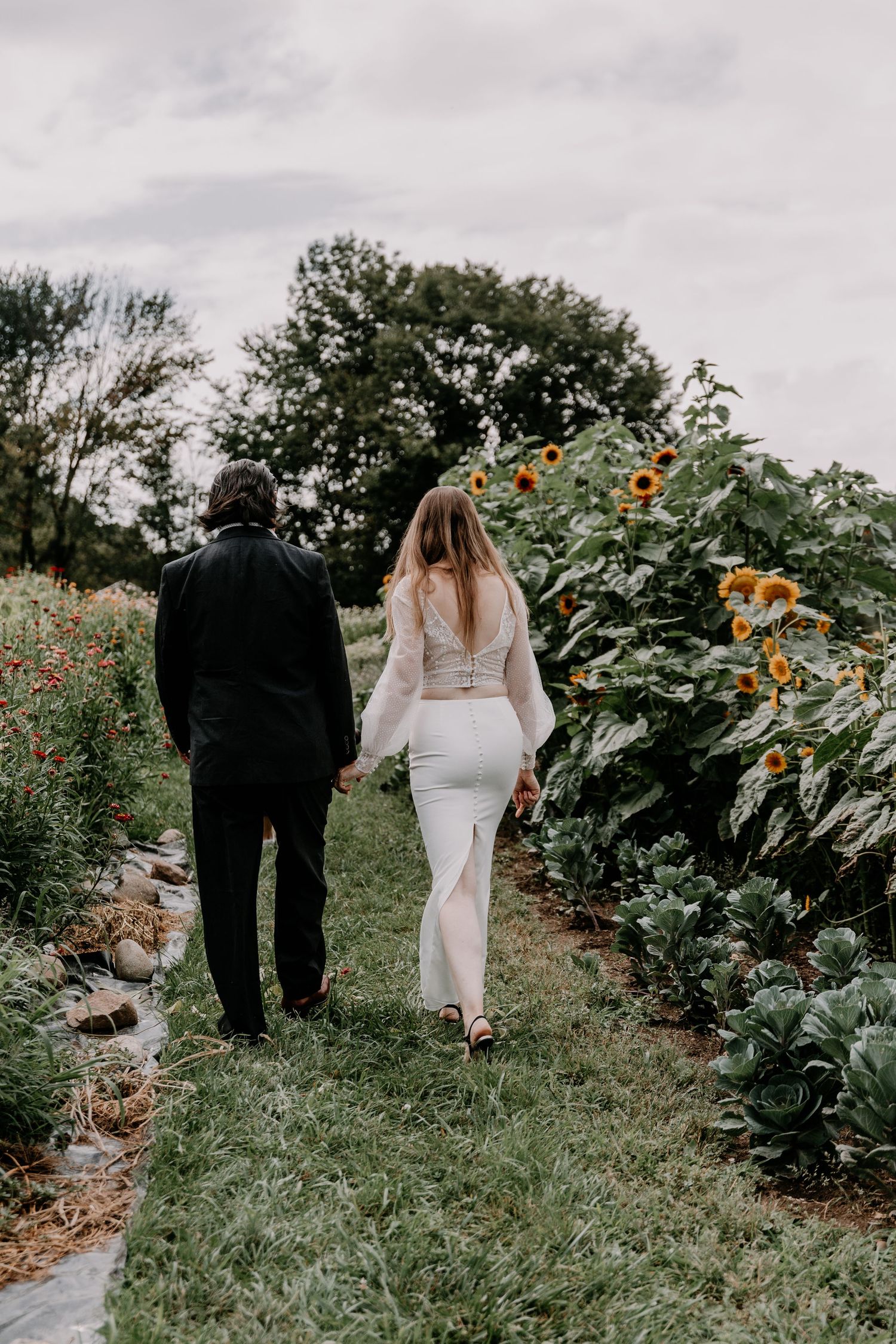 Couple walking down garden path bordered by tall sunflowers and wildflowers on overcast day.