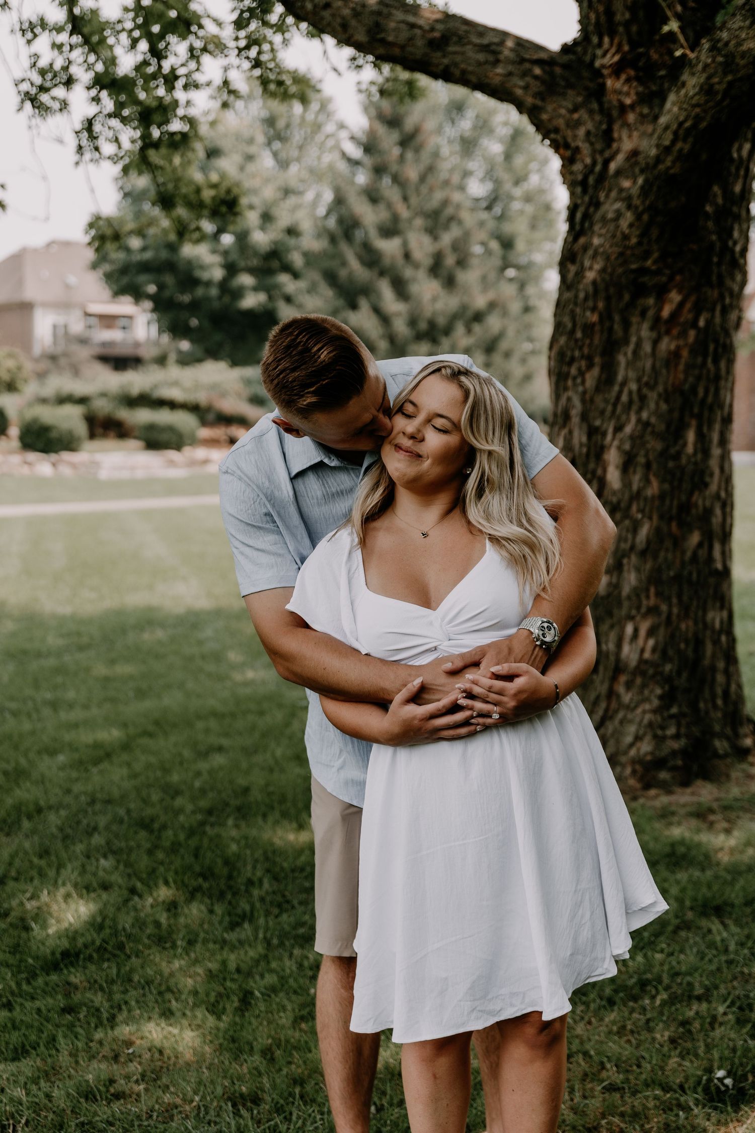 Romantic couple embraces under large tree in park setting during summer photoshoot.