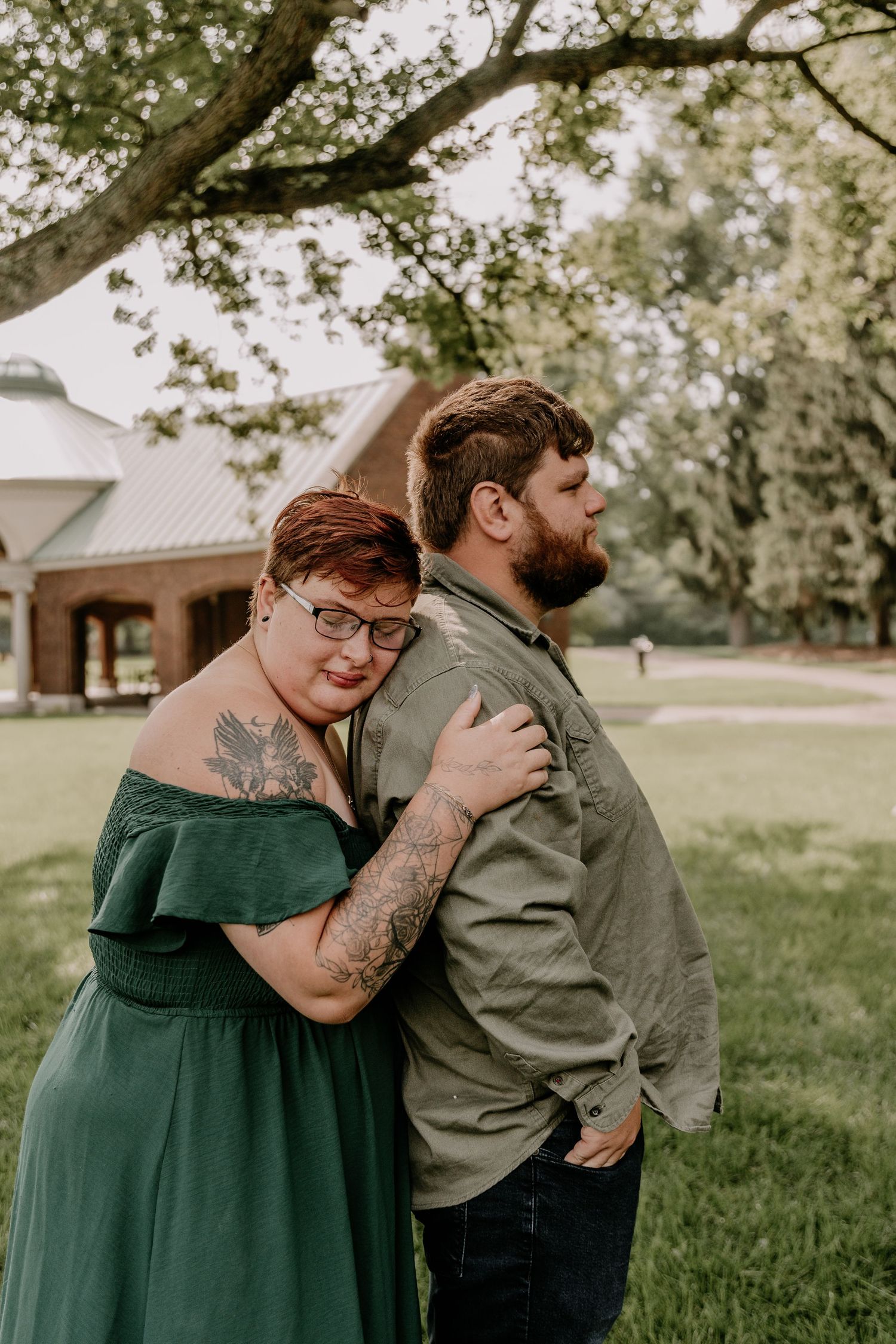 A couple poses back-to-back outdoors, woman in green dress with tattoos leans on bearded man in olive shirt.
