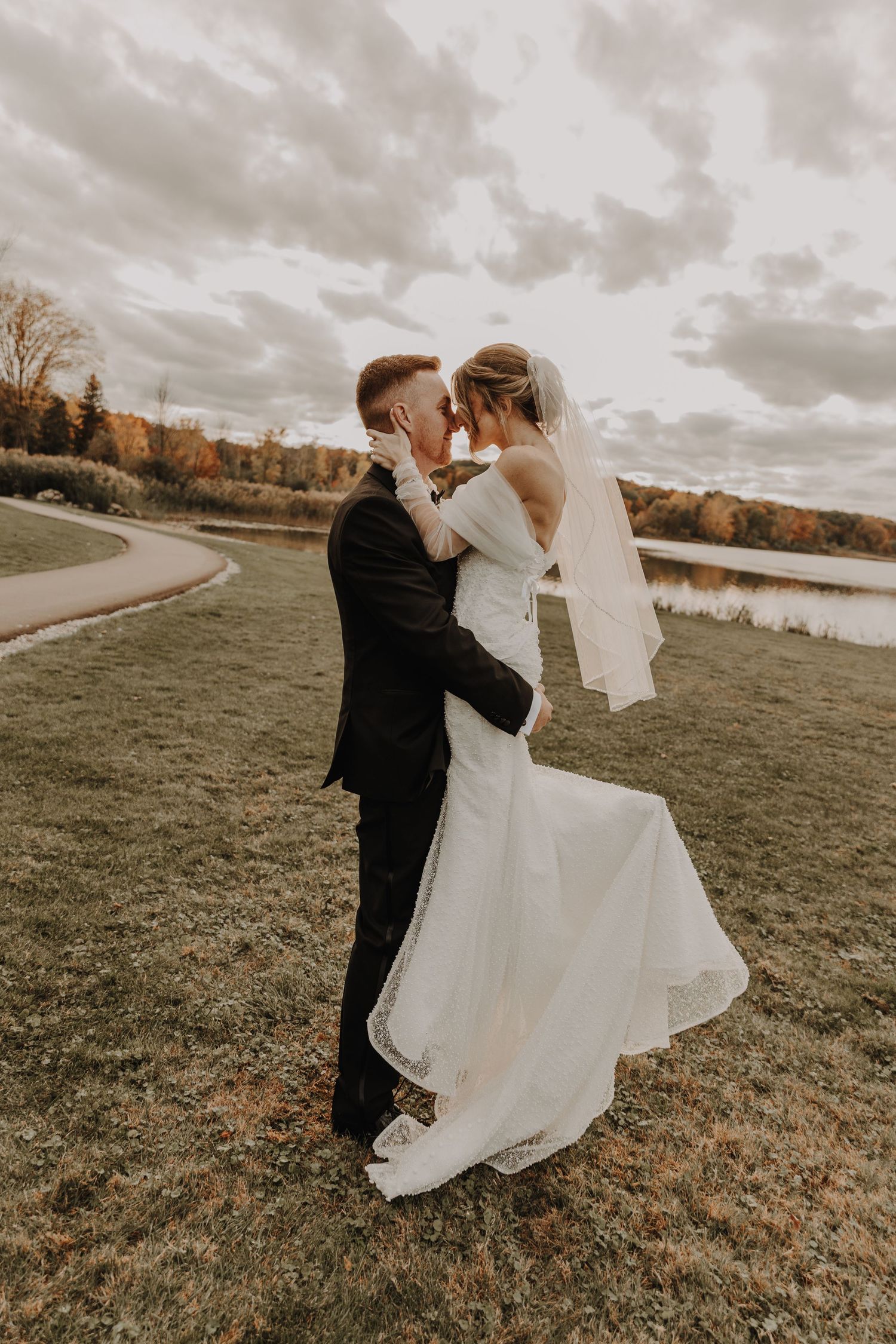 Newlyweds share an intimate moment on a lakeside lawn with fall foliage in the background.