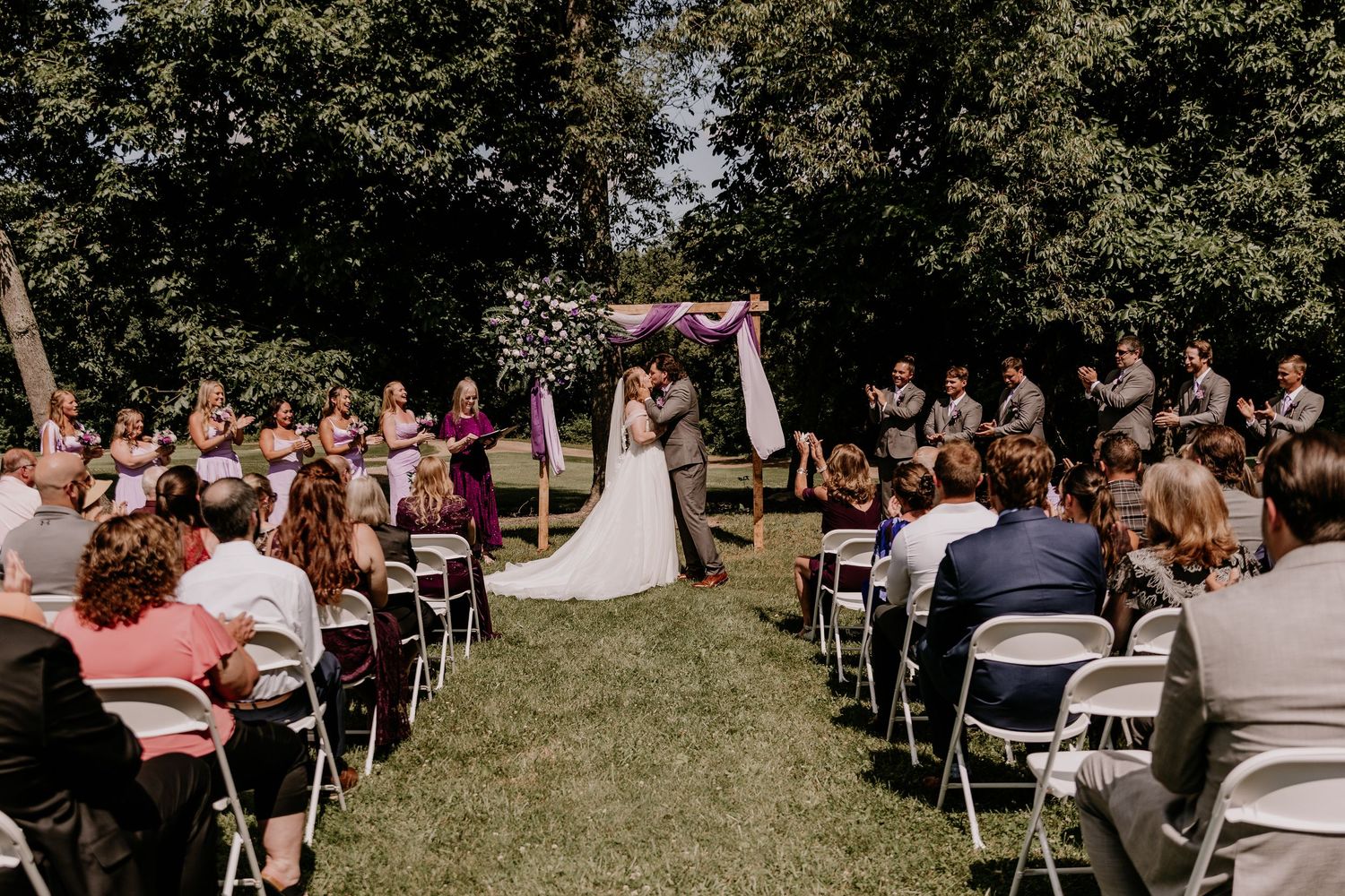Outdoor wedding ceremony with couple at floral arch, guests seated on white chairs on green lawn, surrounded by trees.