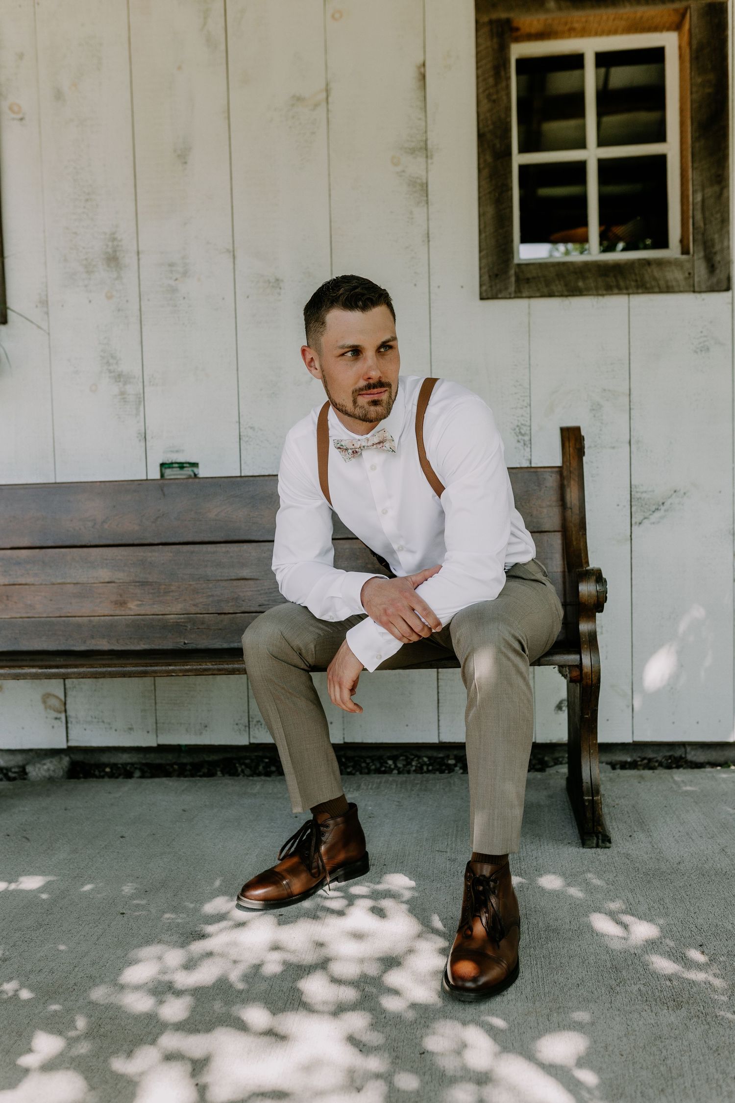 Groom in white shirt, brown suspenders, bow tie, and leather boots sits on wooden bench outside rustic building.