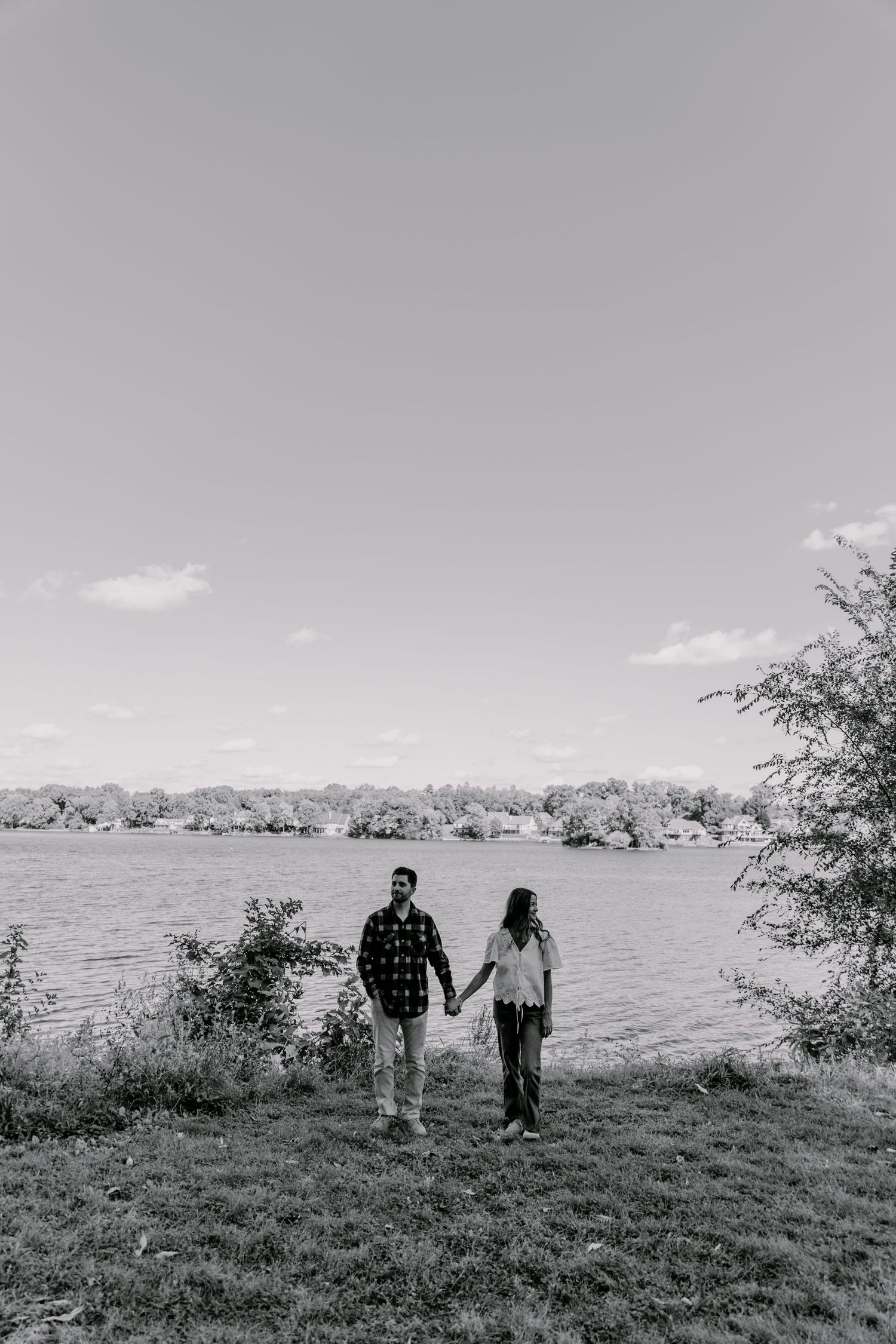 Couple holding hands by a lakeside in black and white photo, surrounded by trees and grass.