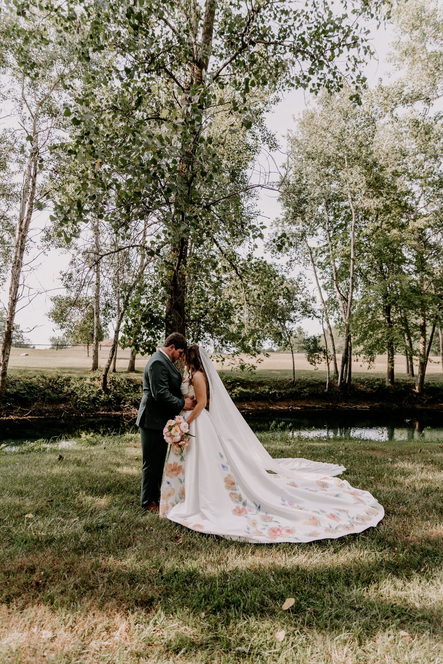 Bride and groom share a romantic kiss outdoors, with a floral-embroidered cathedral wedding dress train spread on green grass.
