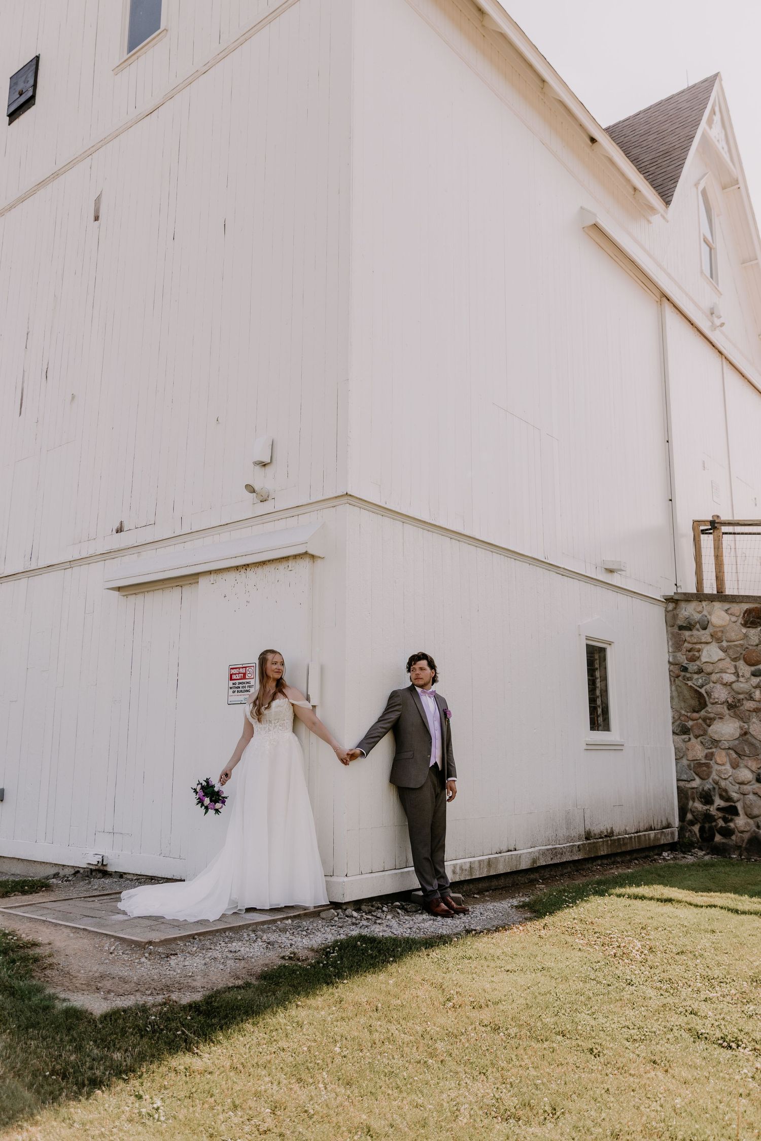 Bride and groom holding hands outside a white barn wedding venue, bride holding a bouquet in a flowing white gown.