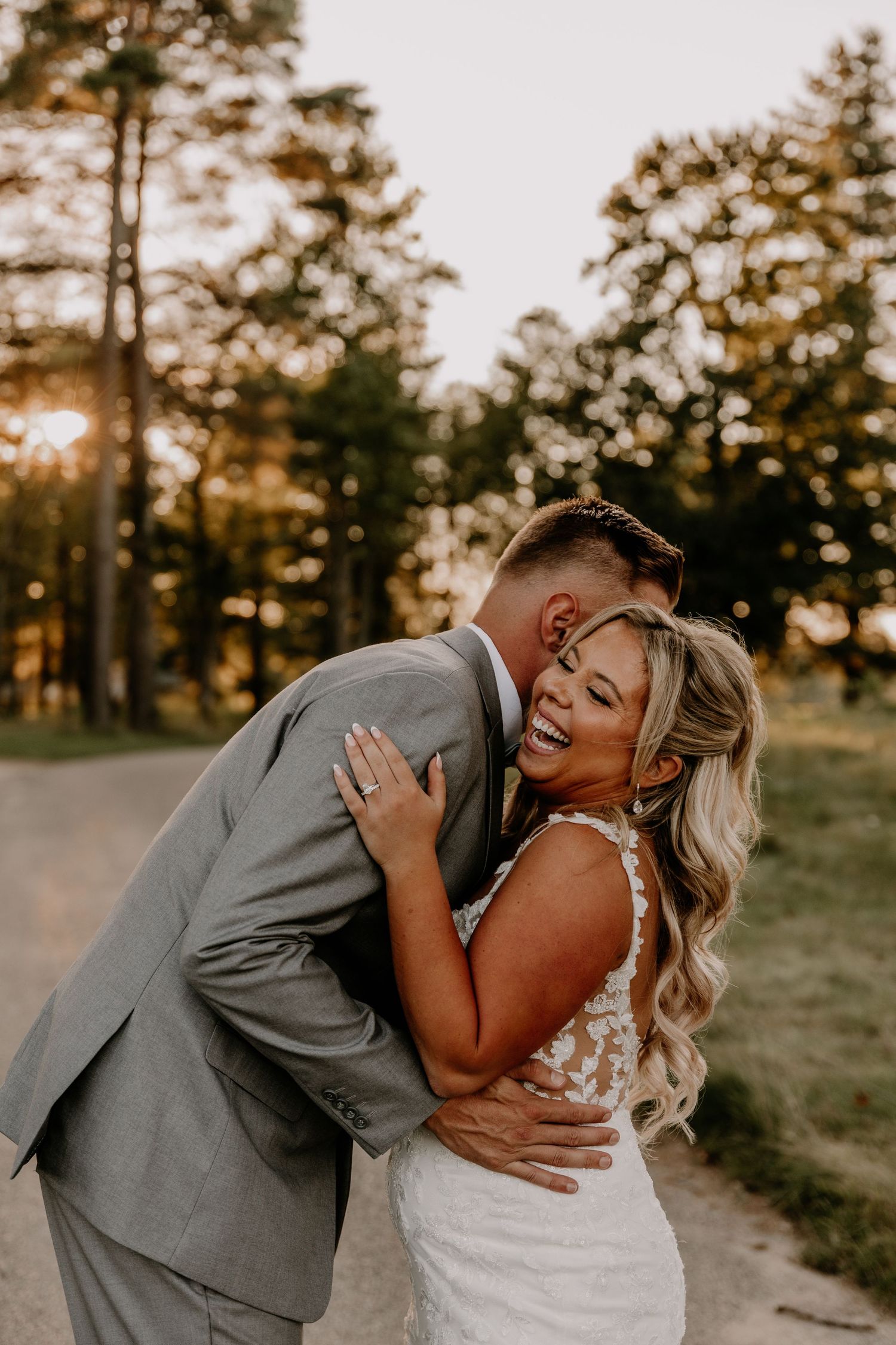 A joyful bride in a lace gown embraces her groom in a gray suit at golden hour outdoors.