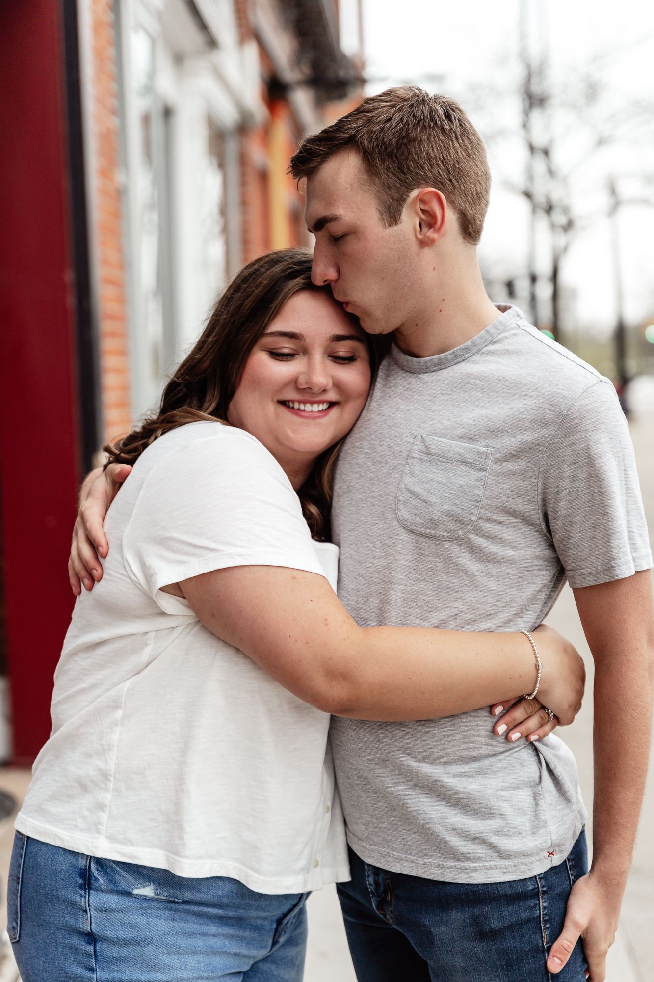 Couple shares romantic embrace against red brick wall during engagement photoshoot.