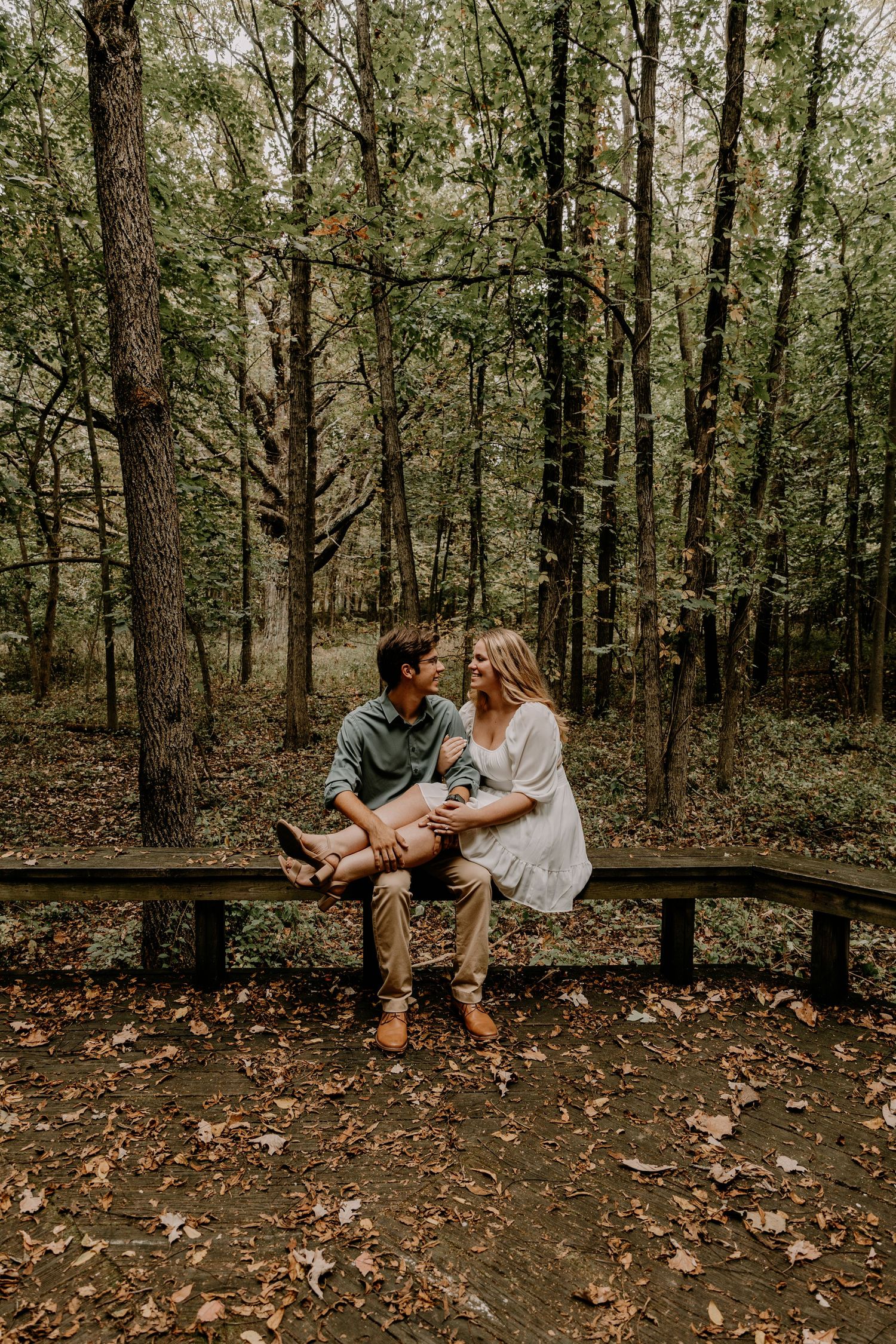 Couple sitting together on wooden bench in wooded forest setting during engagement shoot.