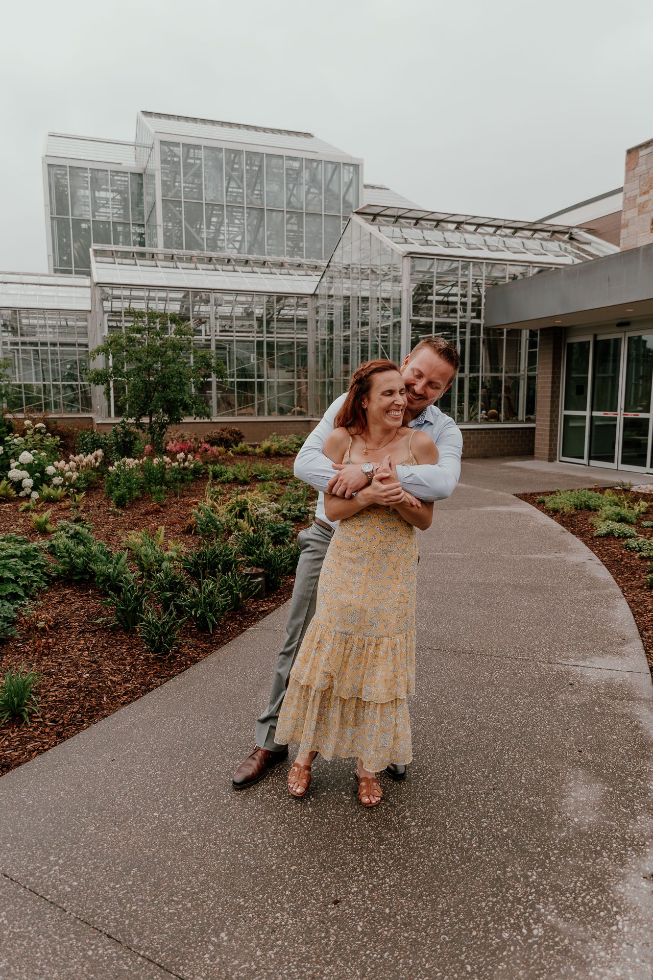 An engaged couple embraces in front of a modern glass building with landscaped gardens.
