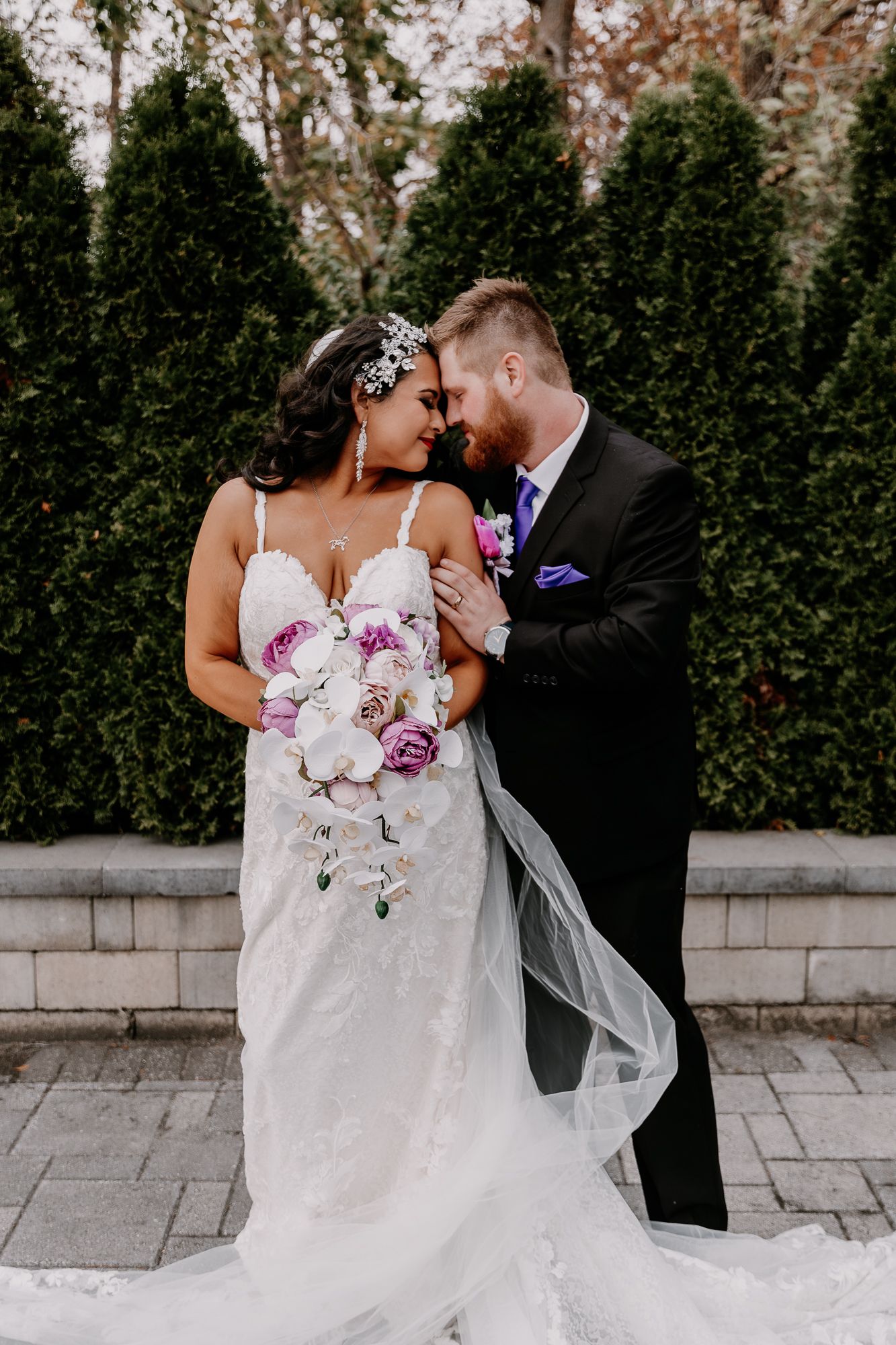 Bride in fitted lace gown holds purple and white bouquet while groom embraces her.