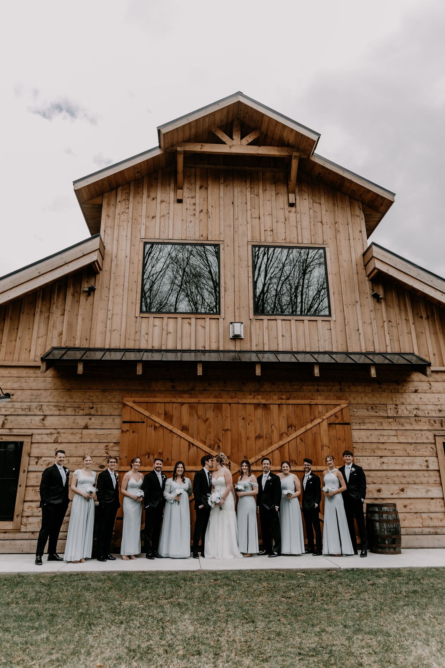 Wedding party poses formally in front of modern rustic barn with wooden exterior and large windows.