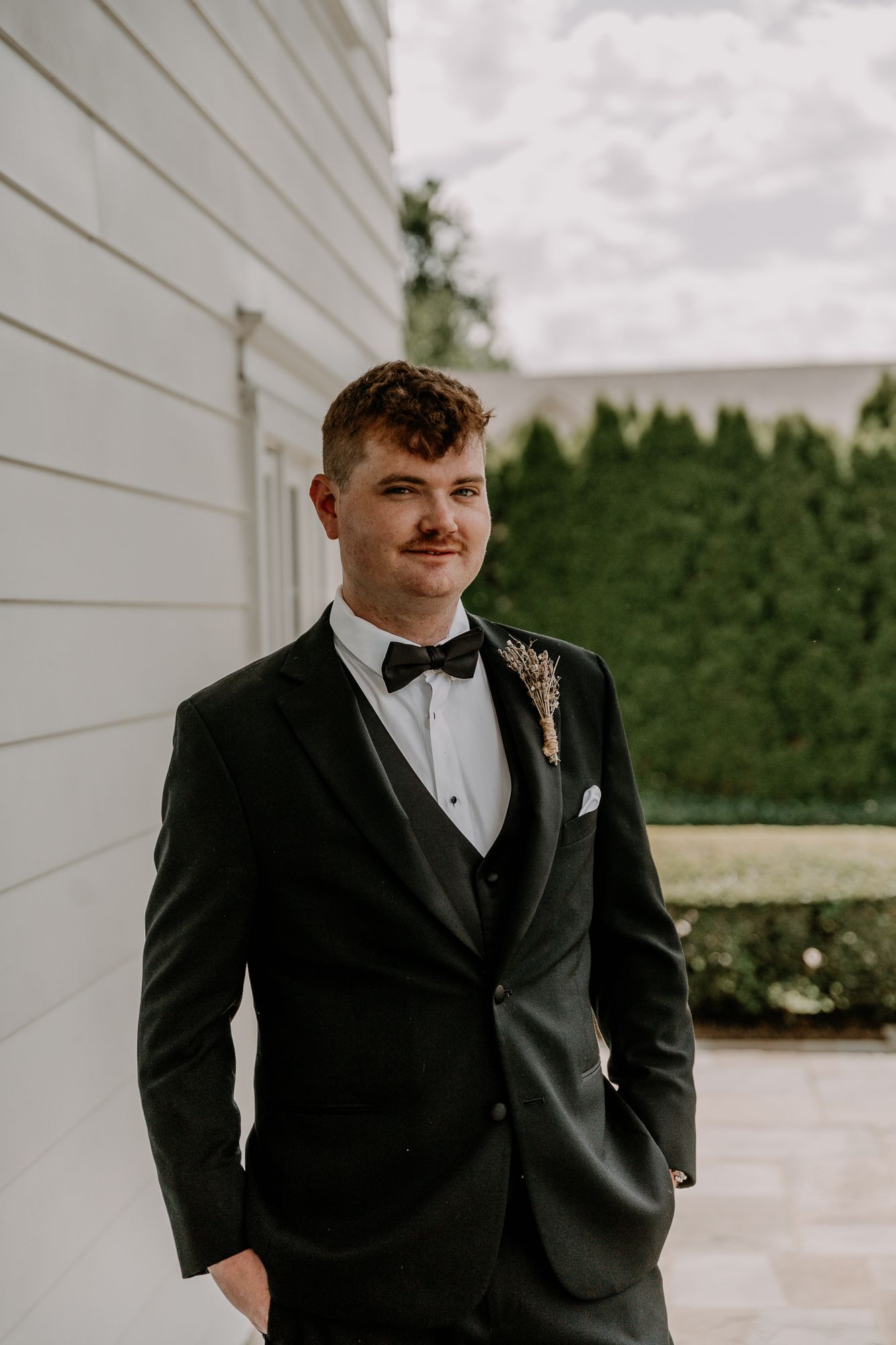 A groom in a classic black tuxedo and bow tie poses against a white building exterior.