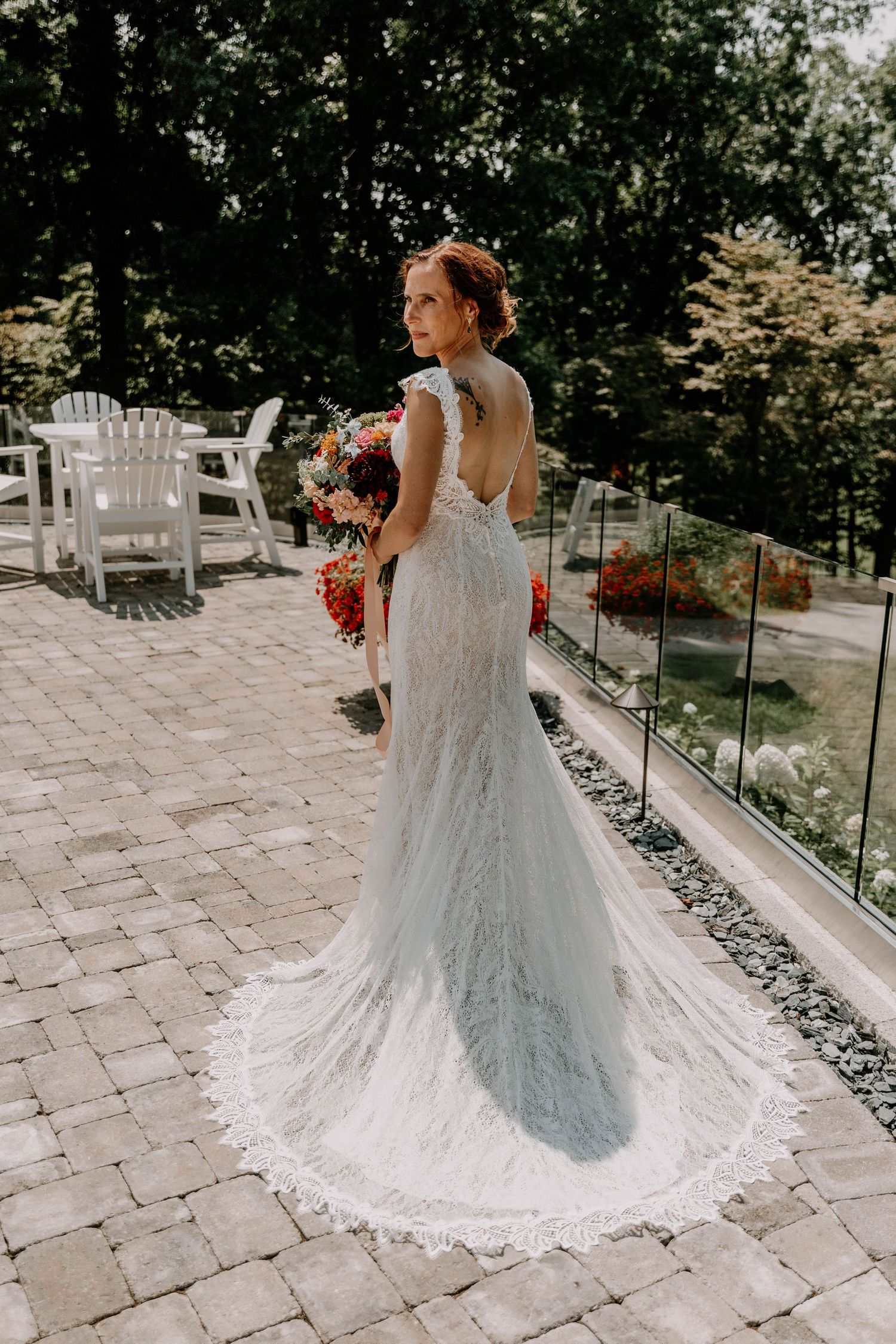 A bride in a fitted lace wedding gown with long train poses on a stone patio with outdoor furniture.