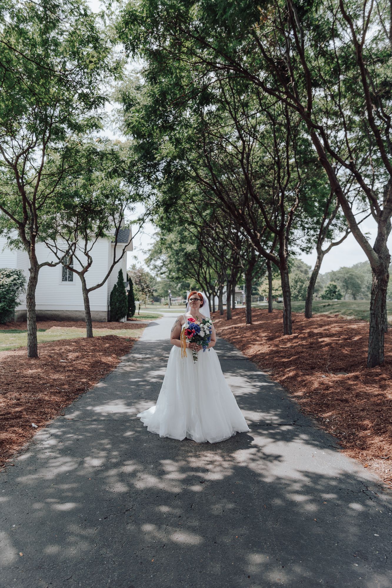 Bride in white gown holds bouquet while standing on tree-lined pathway.