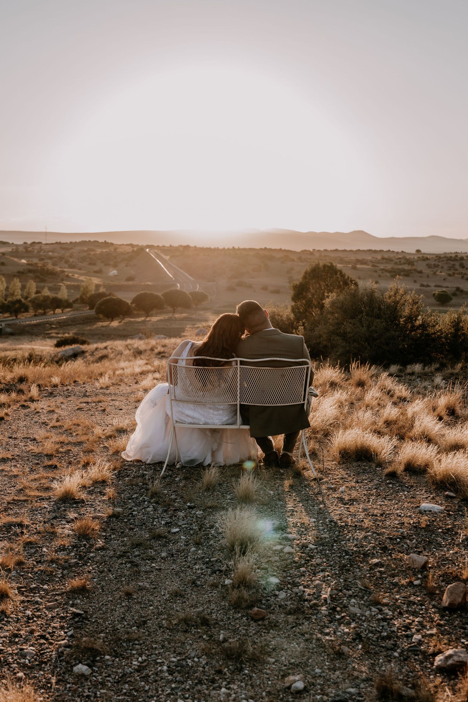 Newlyweds share a tender moment on a vintage couch in a desert setting at sunset.