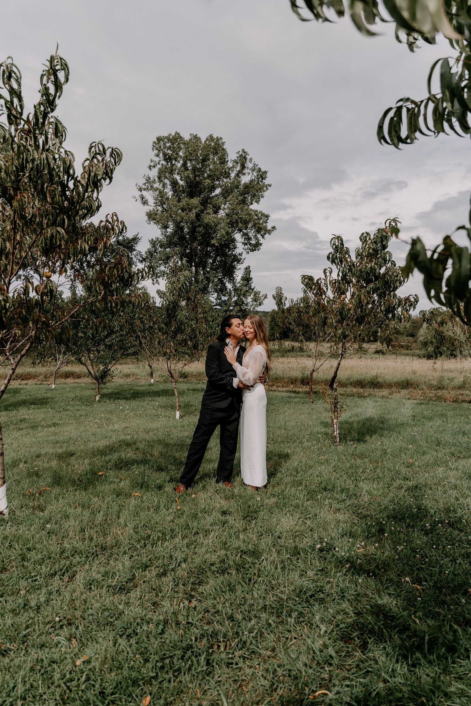Intimate moment between newlyweds in orchard under cloudy sky.