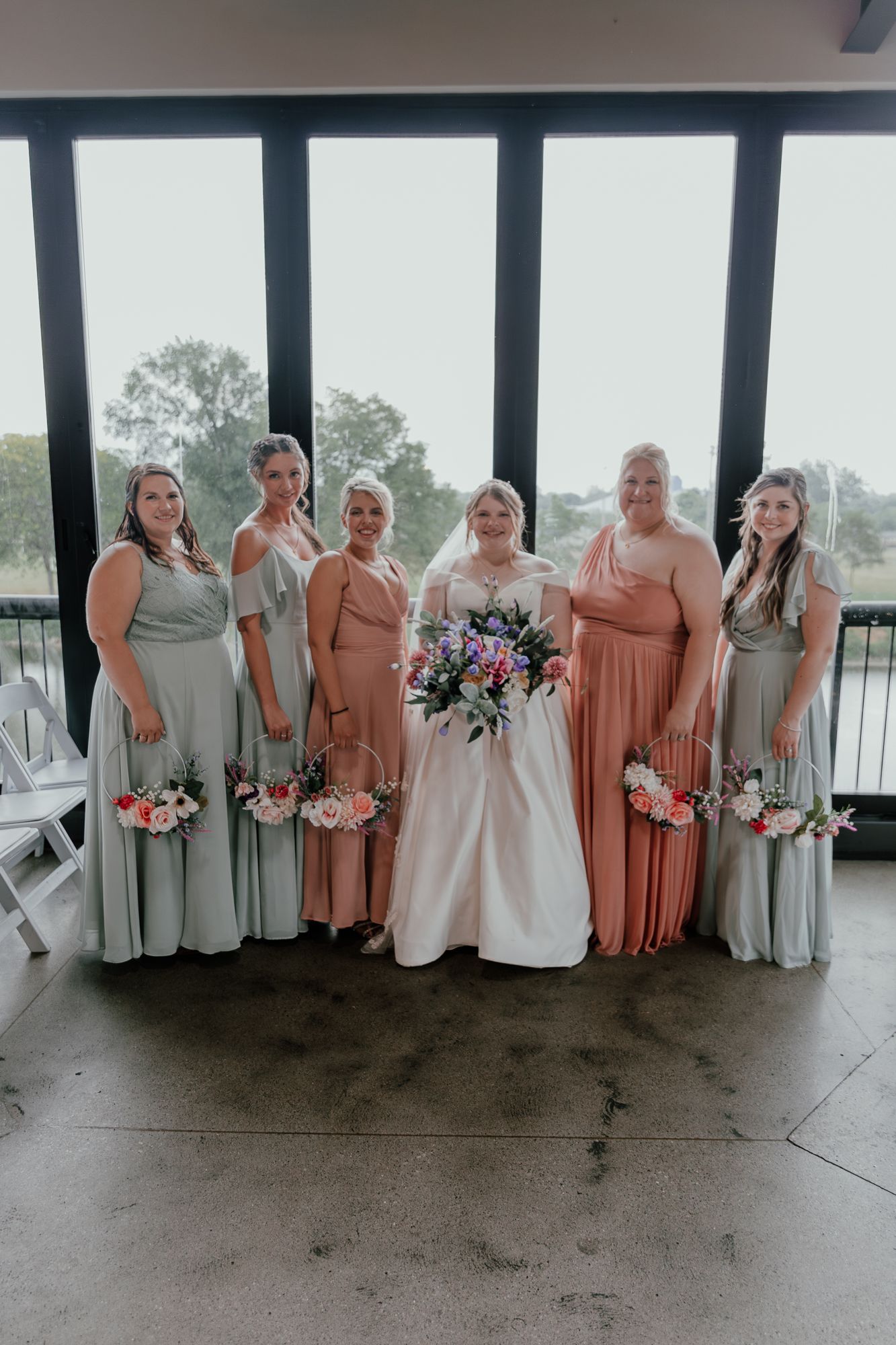 Wedding party in peach and grey dresses poses together in modern venue with waterfront view.