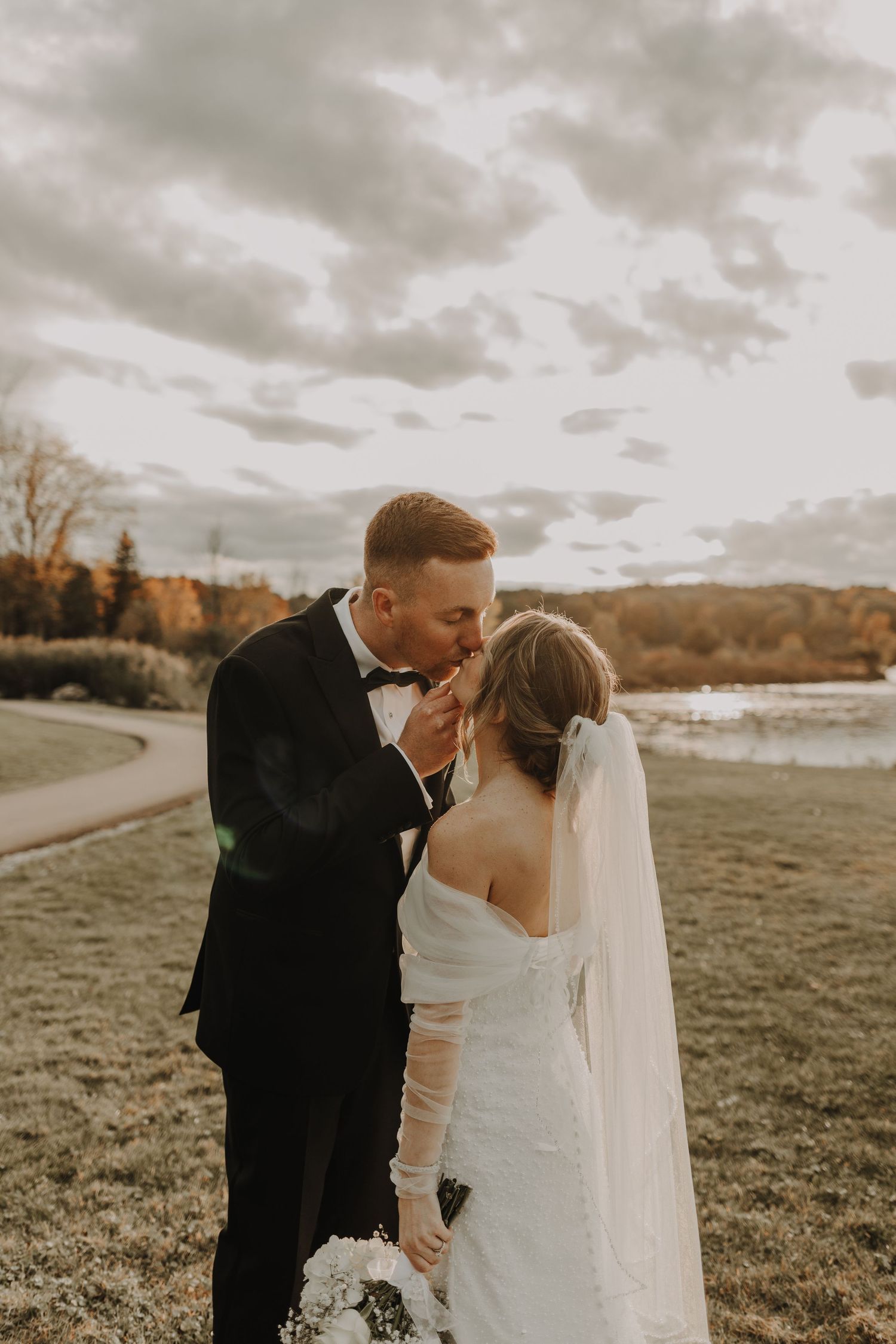 Romantic wedding couple shares intimate moment in autumn field under dramatic cloudy sky.