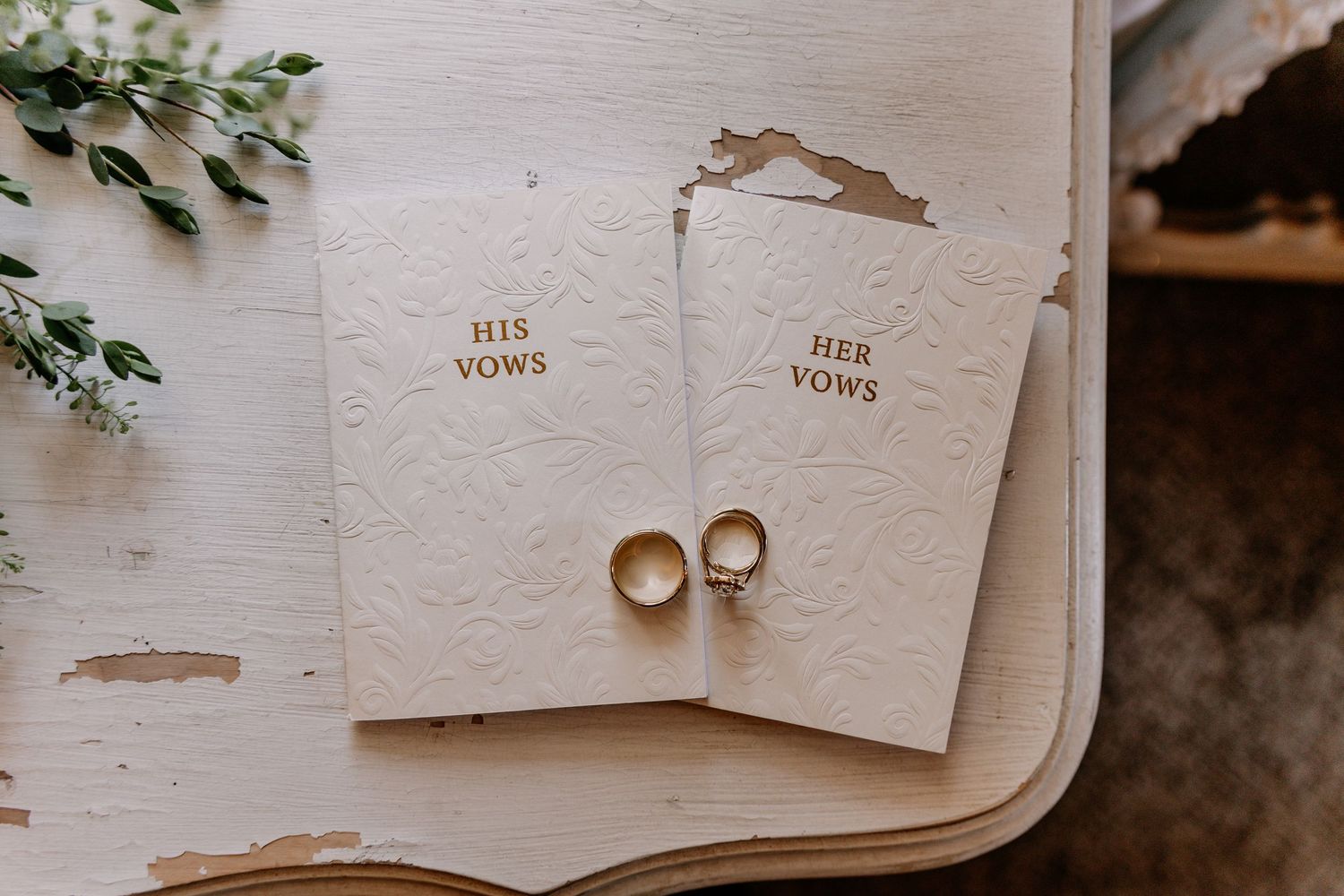 Two gold wedding rings rest on His and Her Vows booklets on a rustic white wooden table with greenery.