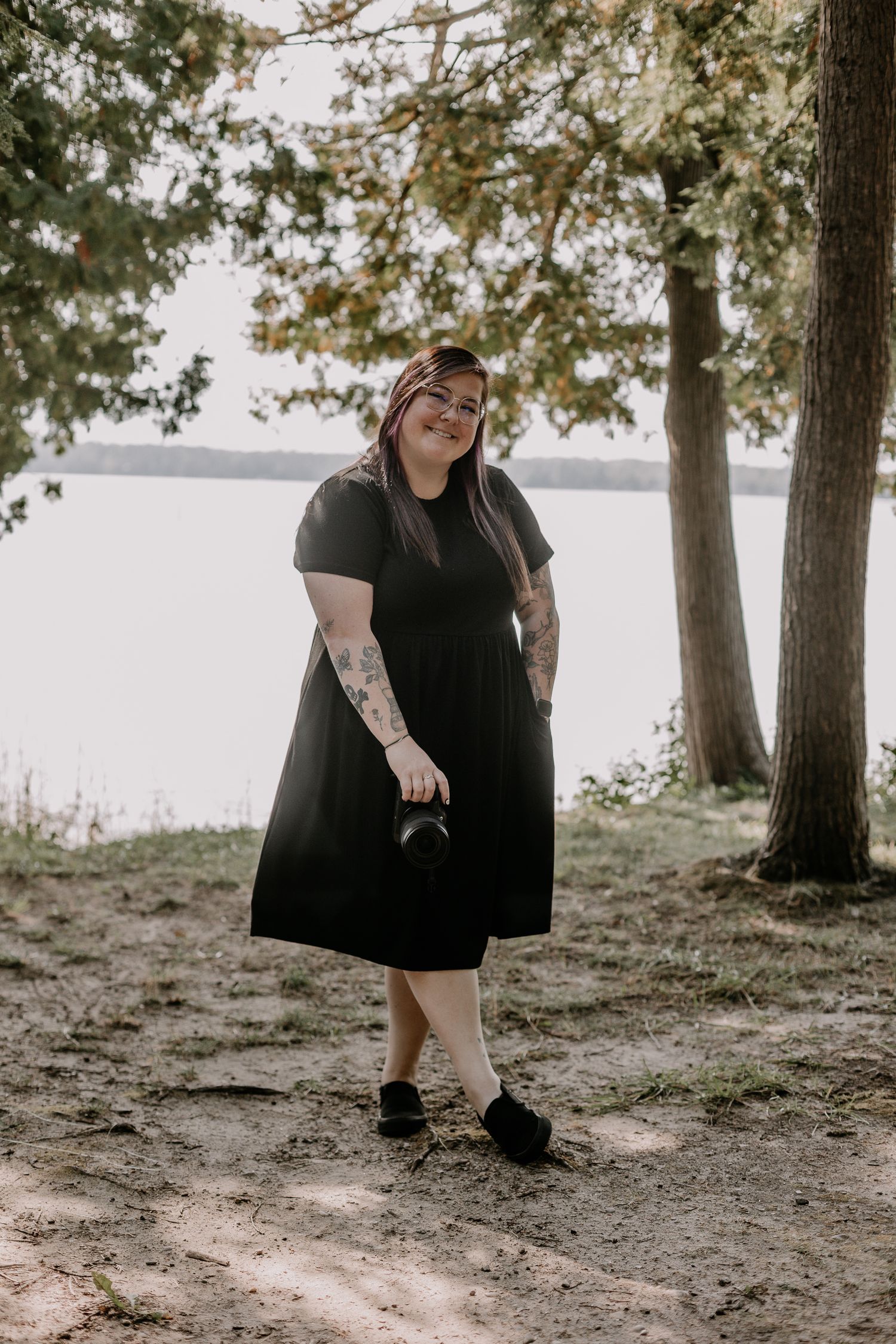 A black-clad figure sits casually on a hillside with tall trees and a lake in the background on an overcast day.
